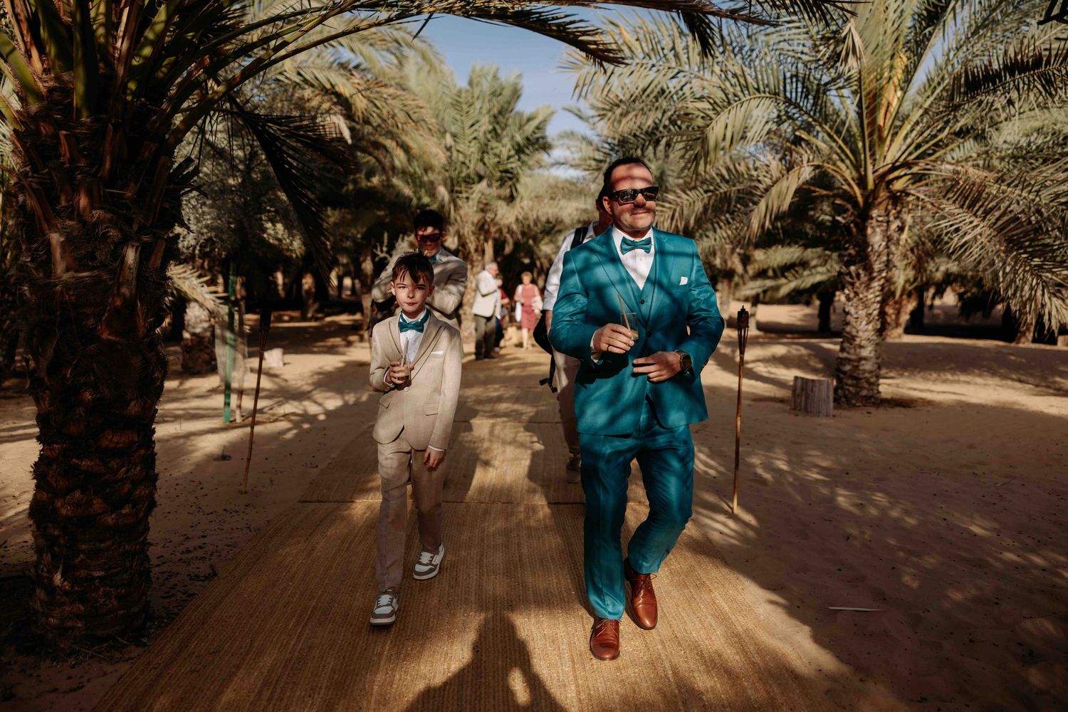 Man in teal suit and child running joyfully through a palm tree-lined sandy pathway in bright sunlight.