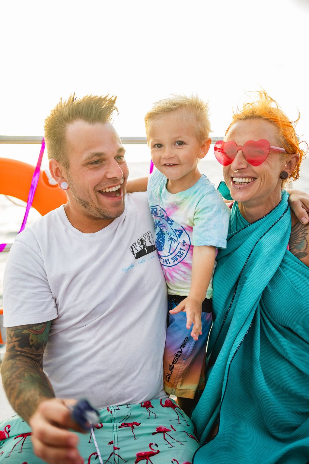 Two tattooed adults laugh while holding a smiling toddler on a colorful boat.