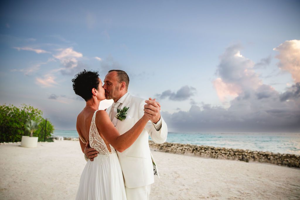 Bride and groom share romantic kiss on tropical beach at sunset with turquoise ocean backdrop.