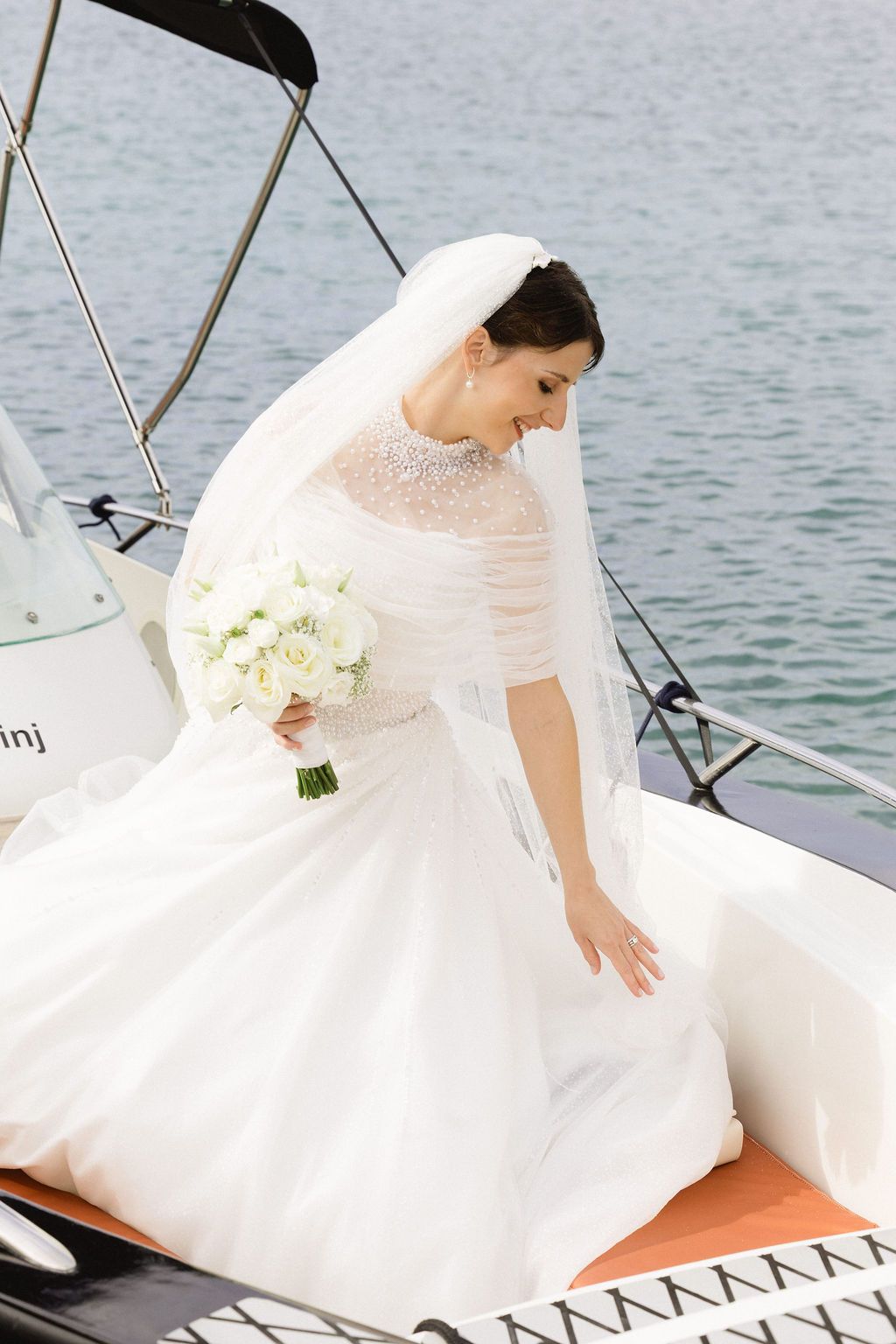 Elegant bride in white ball gown with veil holding white bouquet while seated on yacht.