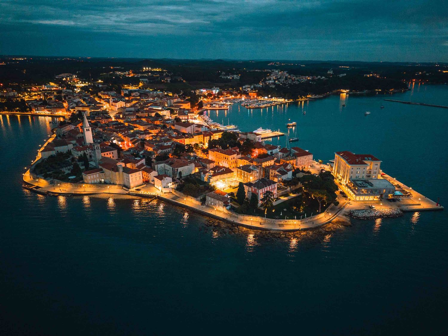 Aerial night view of a illuminated coastal old town on a peninsula surrounded by calm blue waters.