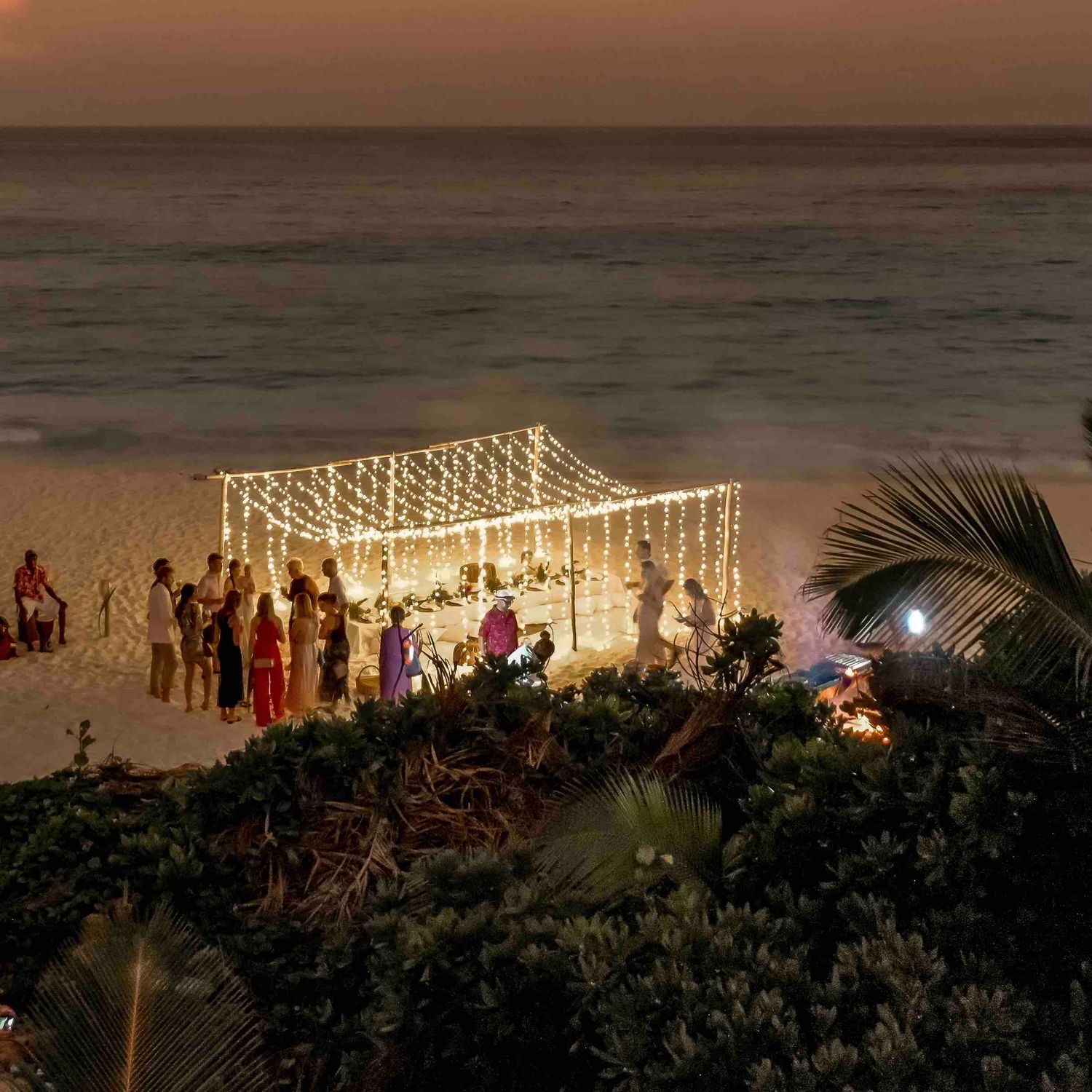 Guests gathered under glowing fairy light canopy on beach at sunset for celebration.