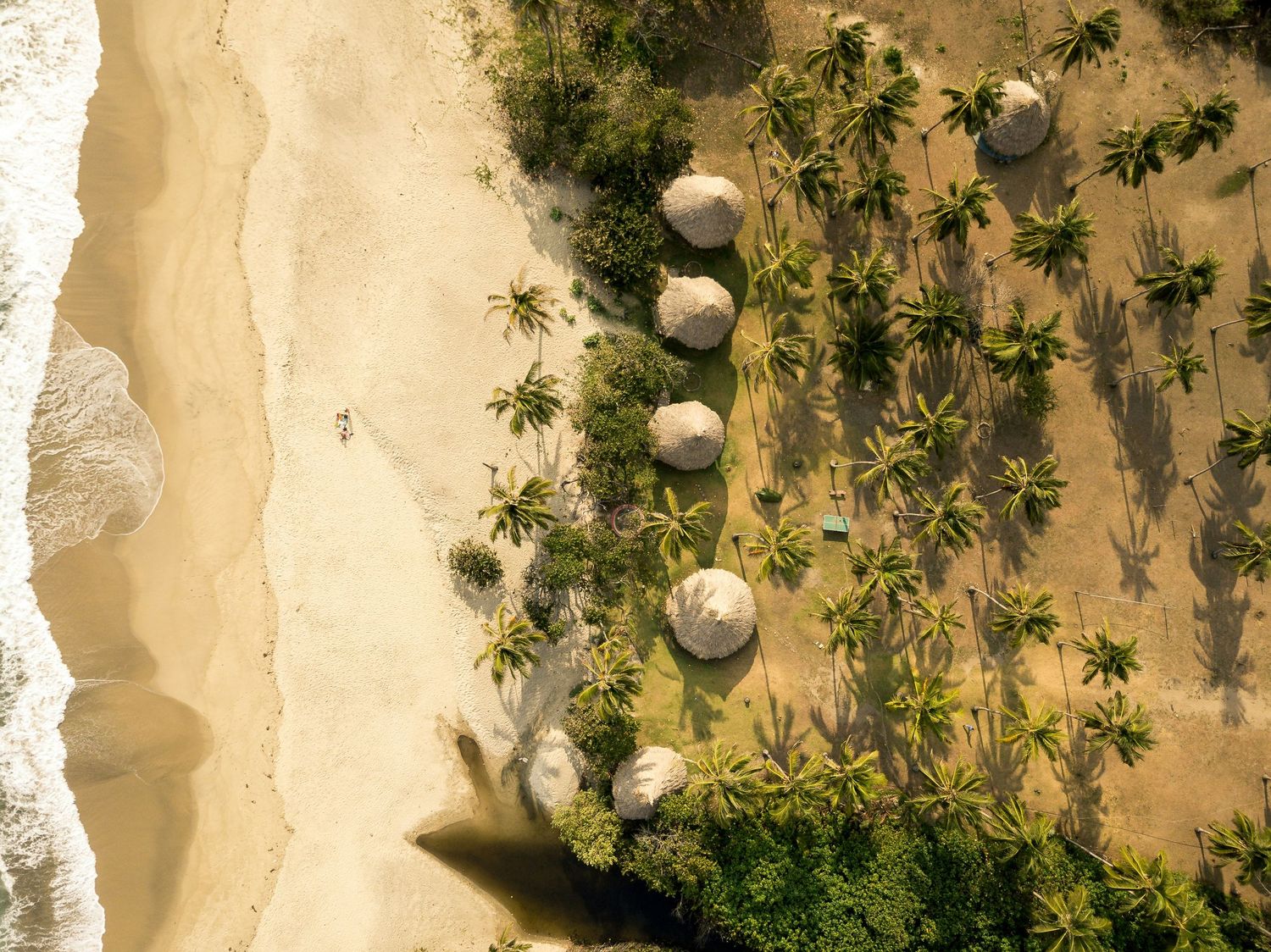 Aerial view of a tropical beach with palm trees, thatched umbrellas, and a lone person walking on golden sand.