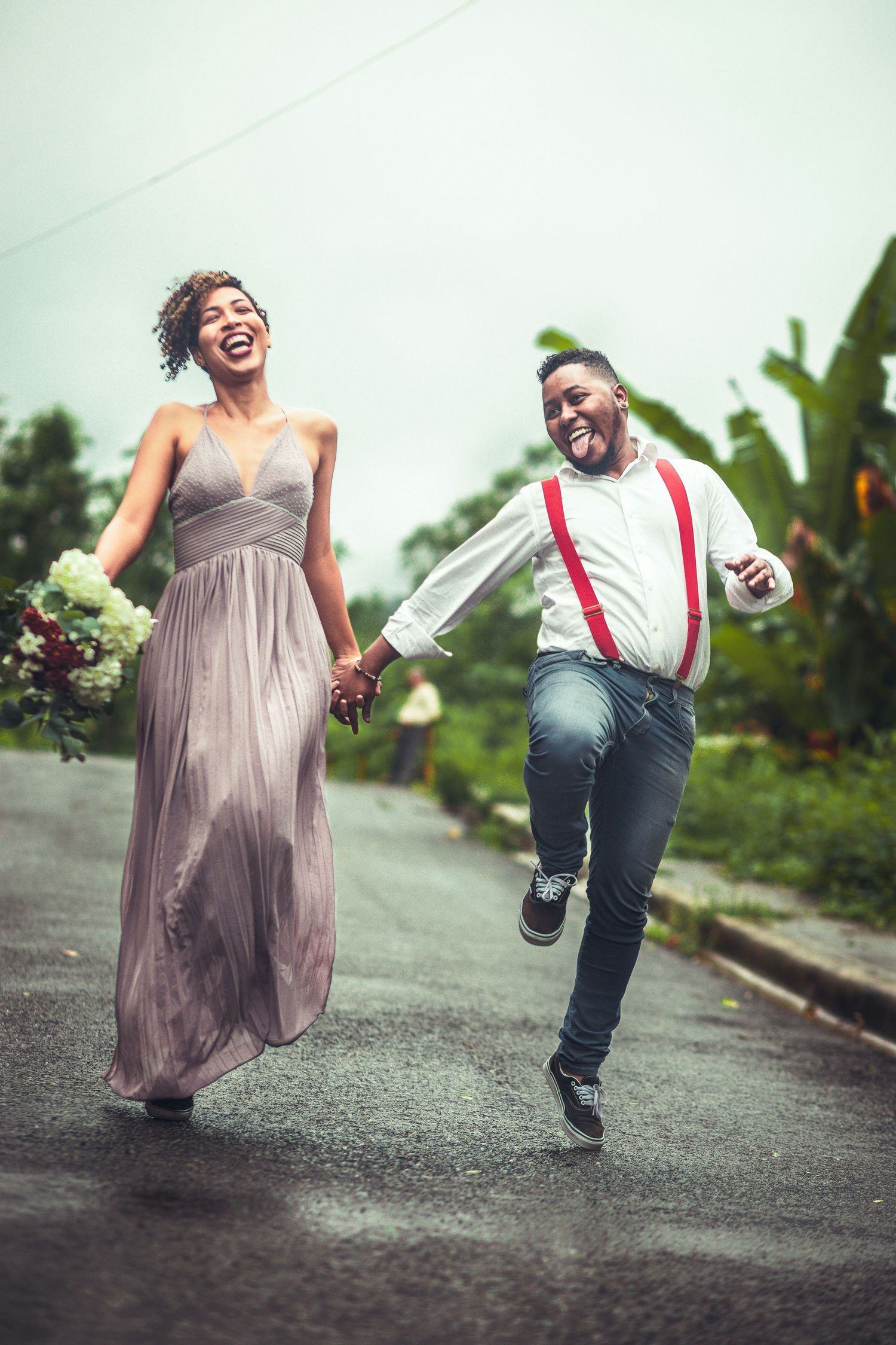 Joyful newlywed couple running and laughing together on rainy road with bouquet.
