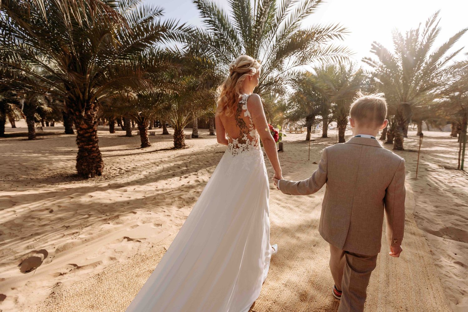 Bride in lace gown and groom in tan suit walk hand-in-hand through a scenic desert palm tree grove.