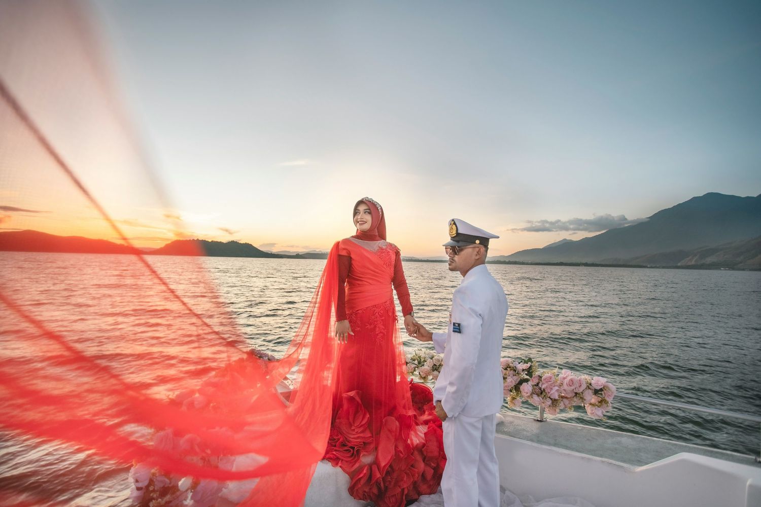 Couple in elegant attire posing on a yacht at sunset with a dramatic red flowing gown.