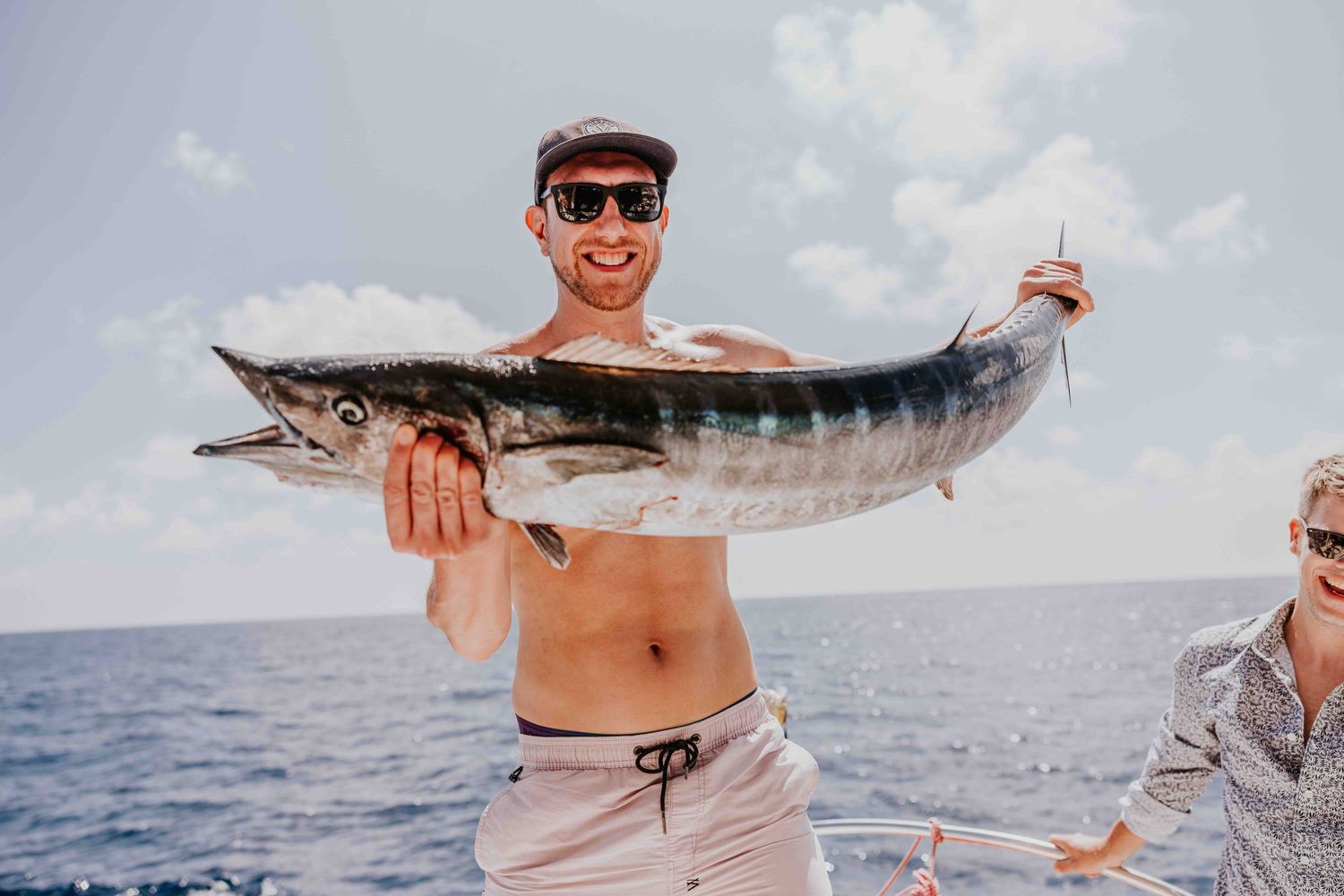 Shirtless man smiling and holding a large wahoo fish on a boat with ocean in background.