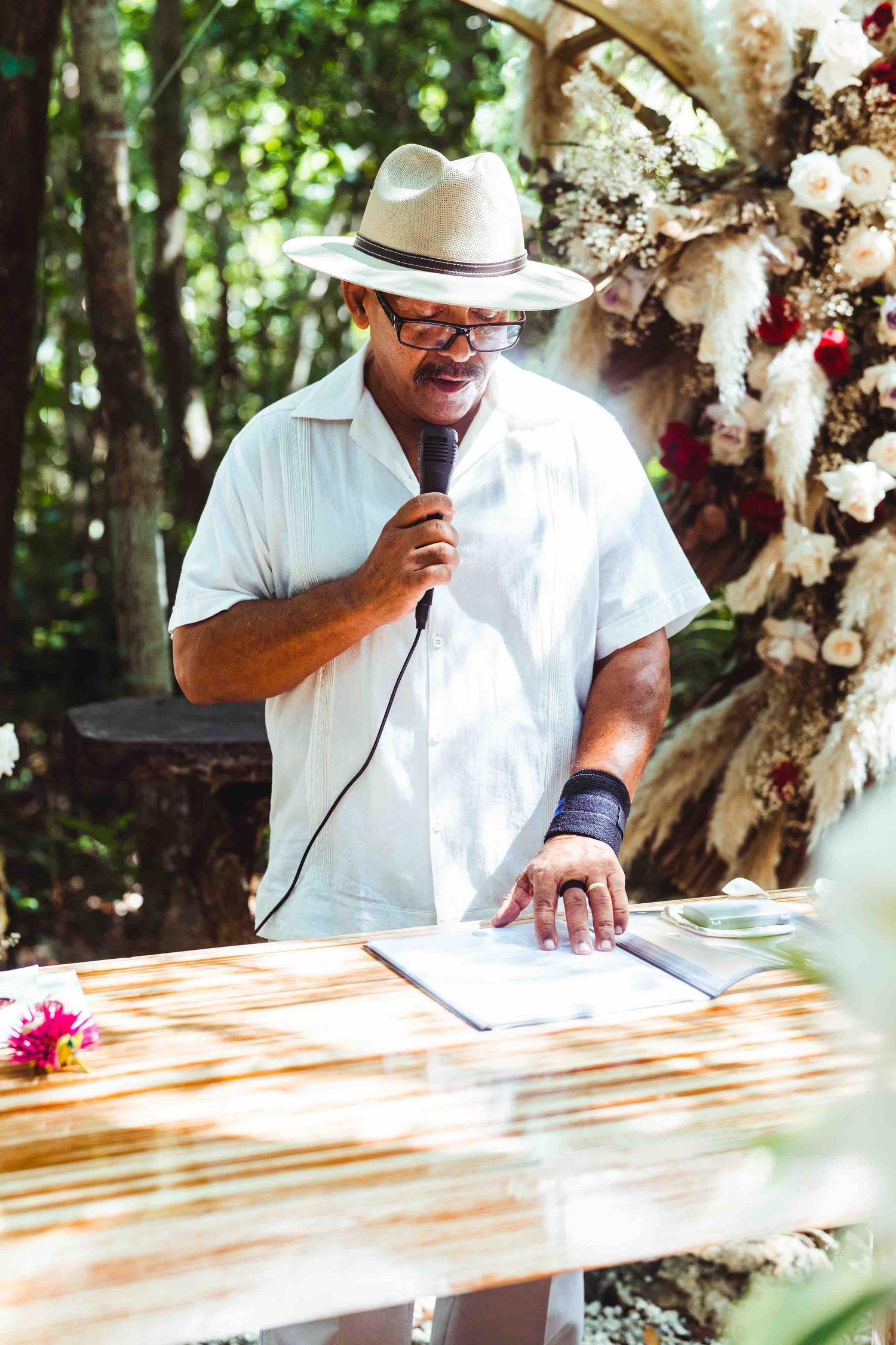 Wedding officiant in white shirt and panama hat speaking into microphone at outdoor ceremony altar.