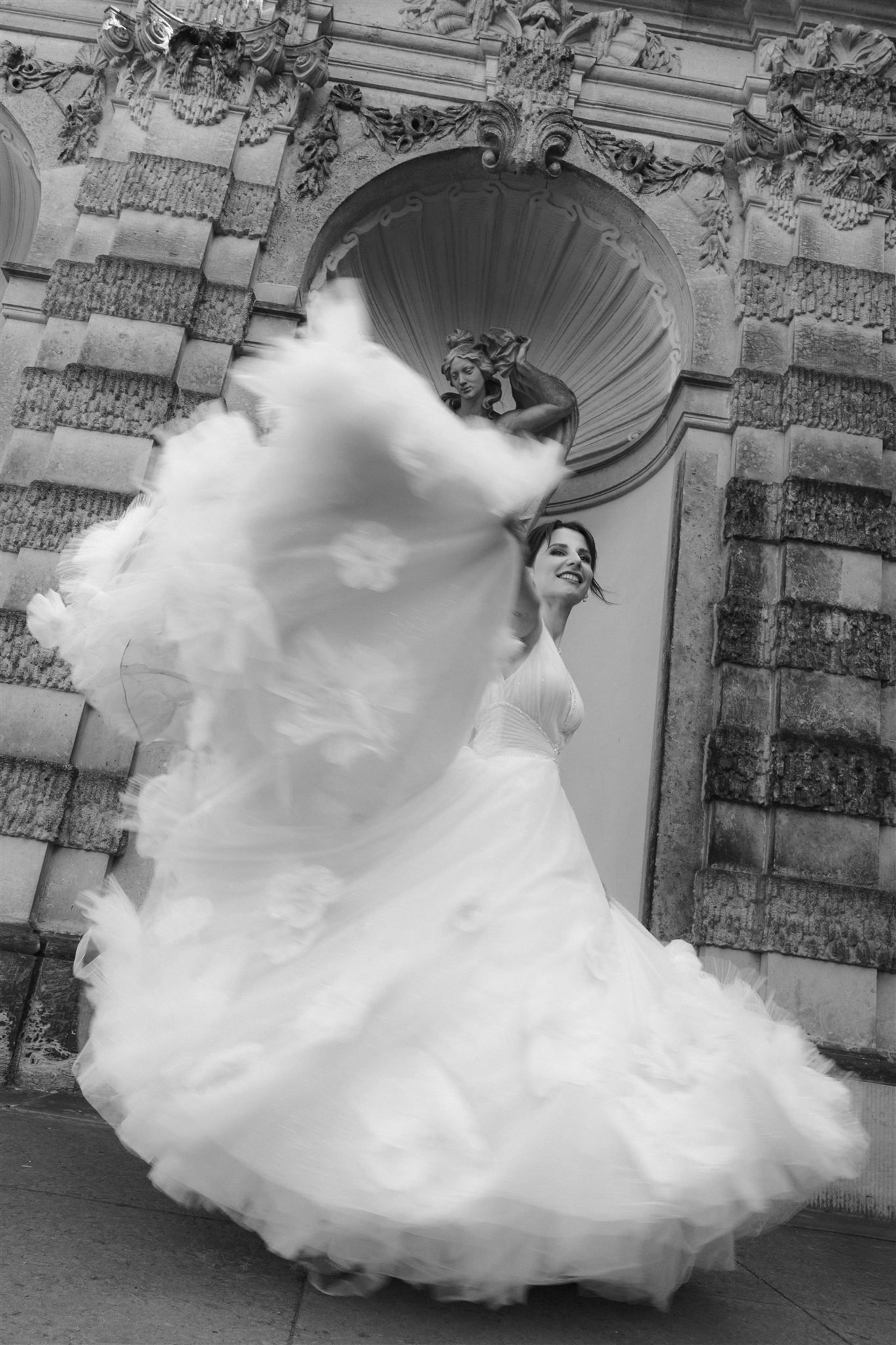 Bride twirling in a voluminous white wedding gown outside an ornate stone building, black and white photo.