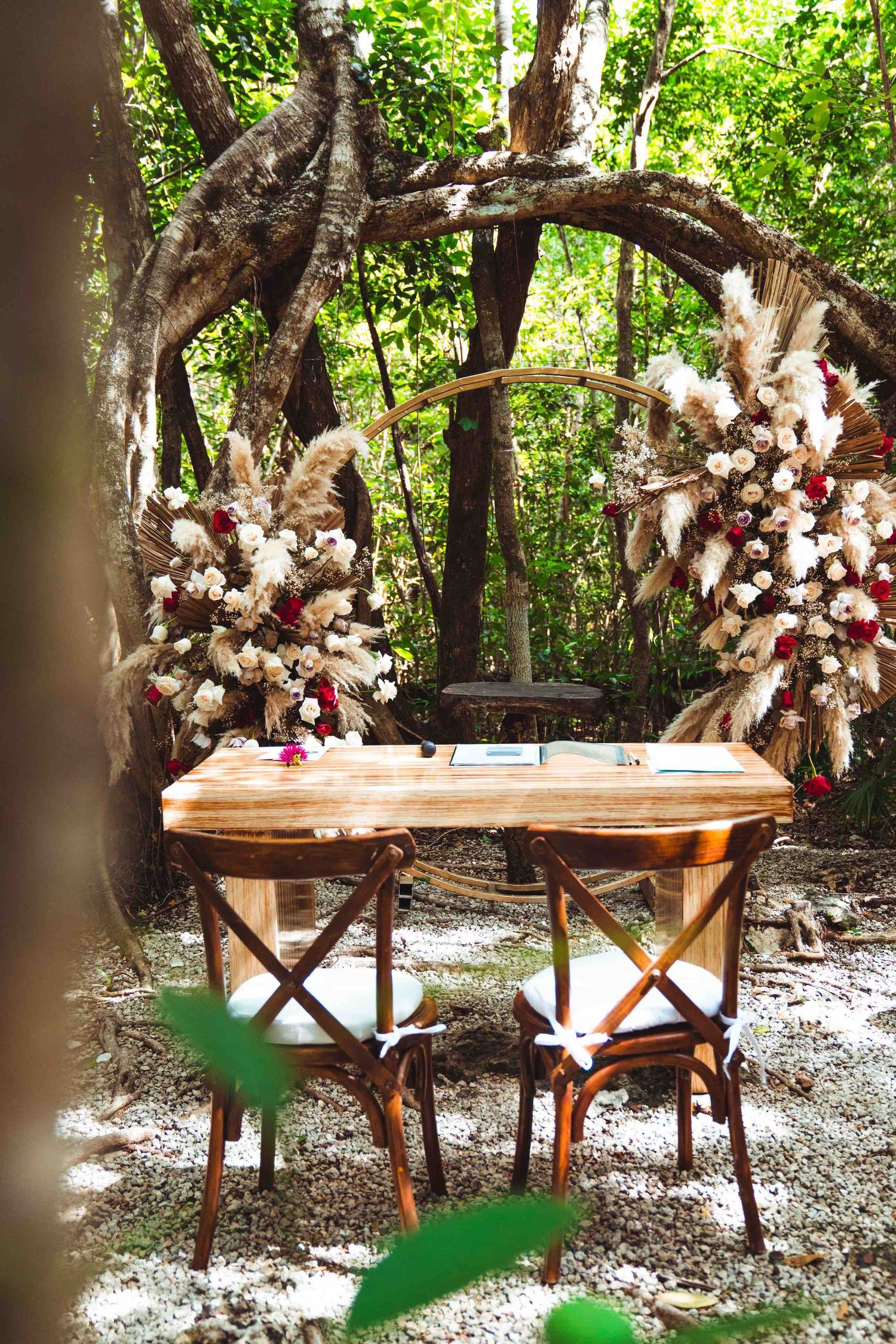 Rustic wooden altar table with cross-back chairs and pampas grass floral arch in forest setting.