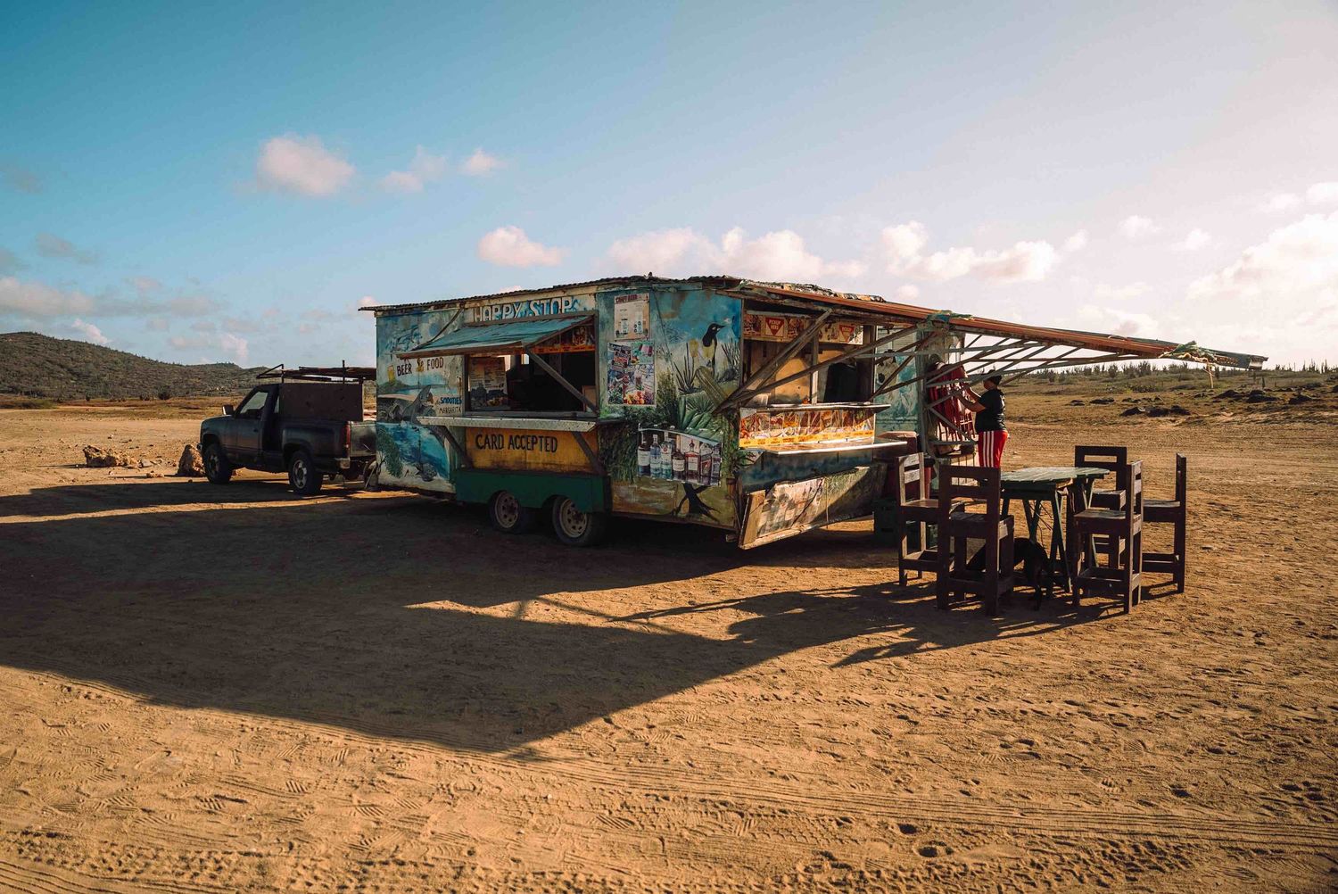 Colorful food truck with outdoor seating on a sandy beach under a blue sky in a tropical coastal setting.
