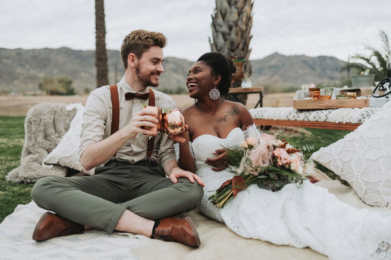 Interracial bride and groom toast with drinks during a romantic outdoor desert picnic wedding celebration.