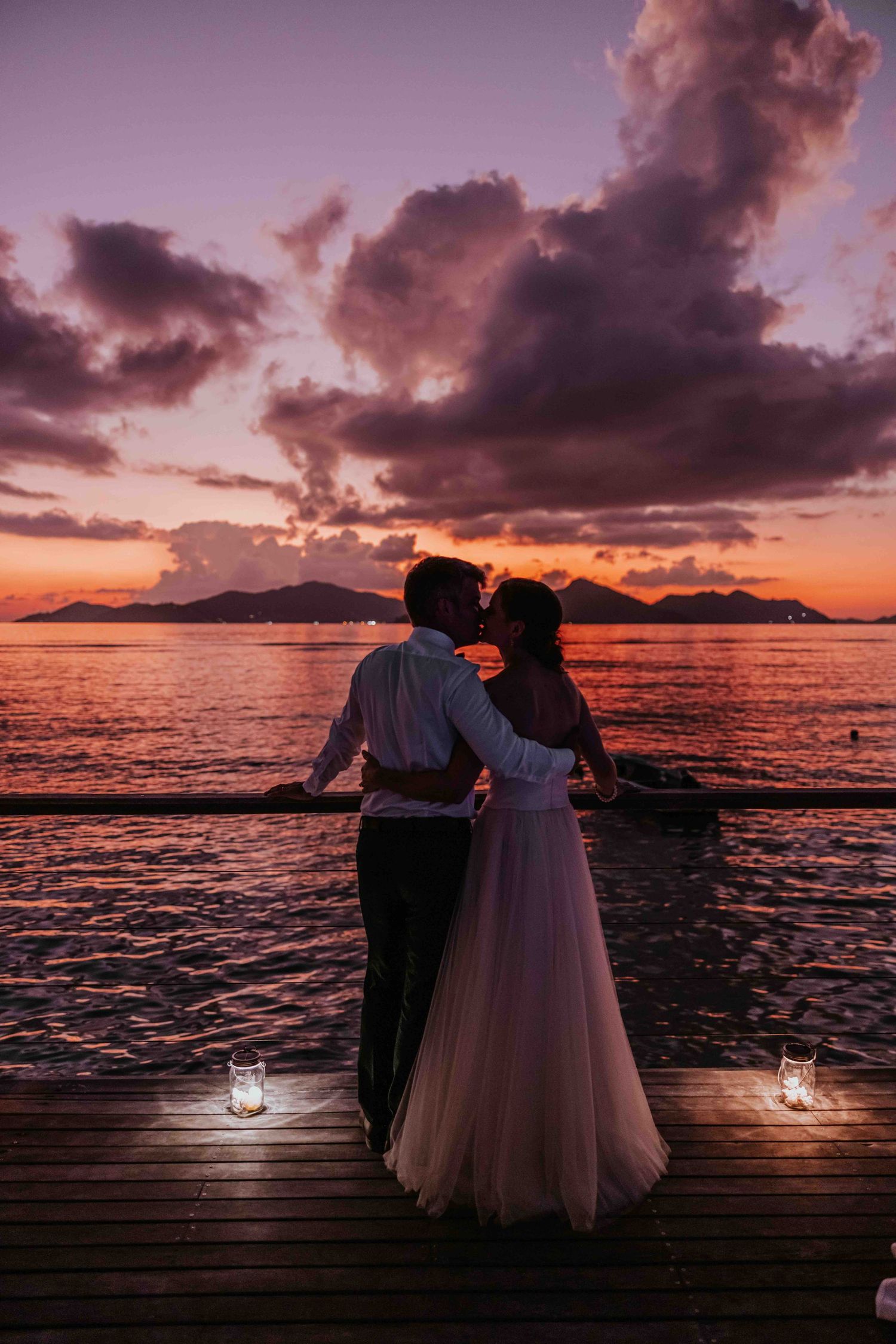 Bride and groom share a kiss on a wooden dock during a stunning purple and orange ocean sunset.