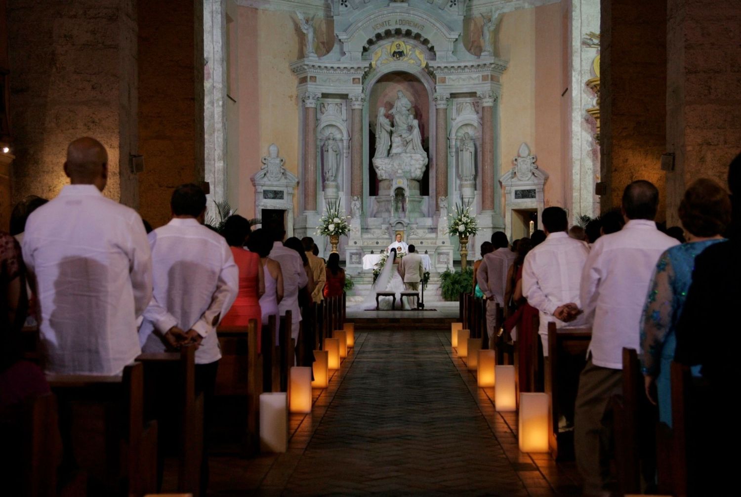 Candlelit church wedding ceremony with guests standing in pews facing a decorated white altar.