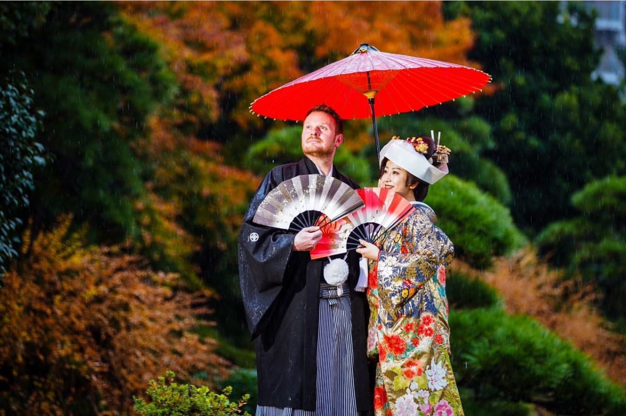 Couple in traditional Japanese wedding attire sharing a red umbrella in autumn garden.