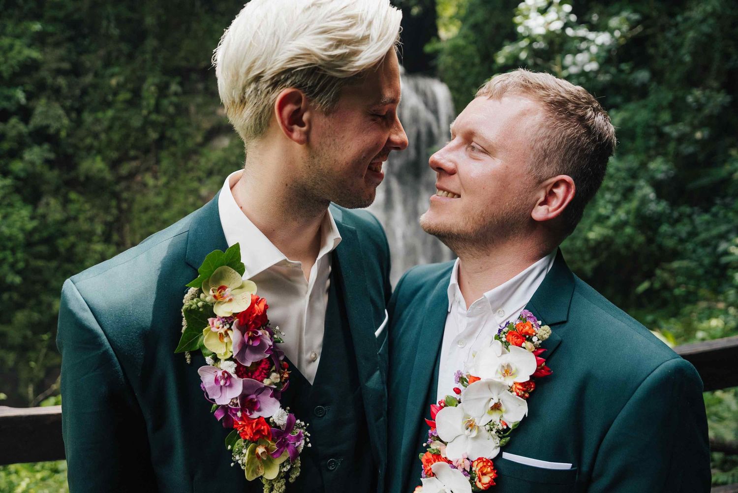 Two grooms in green suits with floral leis smiling at each other outdoors near a waterfall.