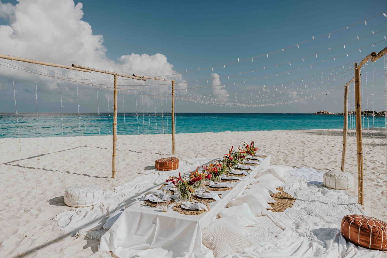 Elegant beach dining table setup with white linens and floral centerpiece on white sand.
