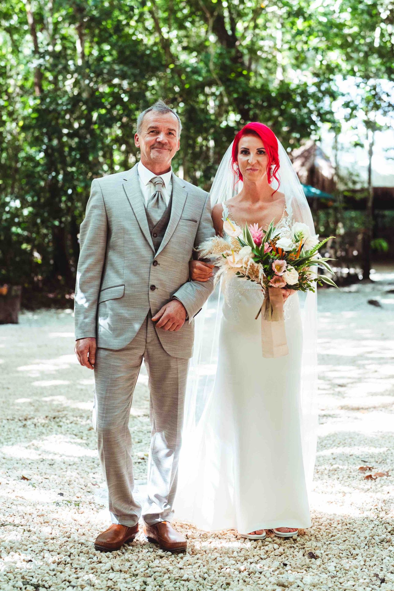 Bride with red hair holding tropical bouquet stands with man in grey suit at outdoor wedding.