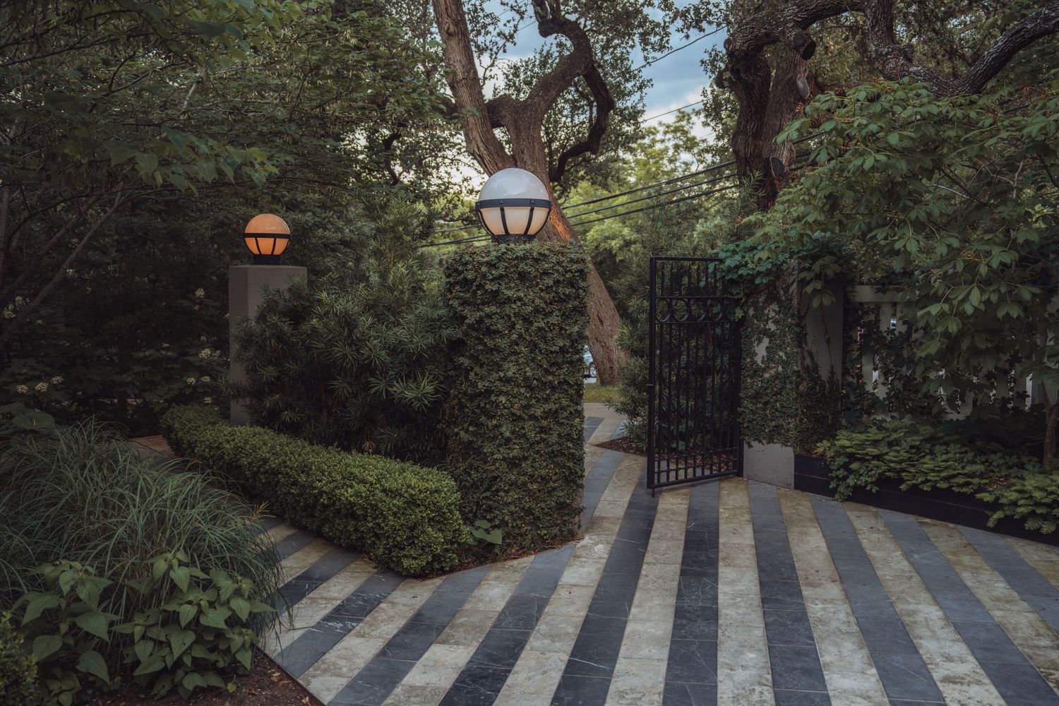 A charming garden entrance with black iron gates, striped stone pathway, and glowing lanterns mounted on hedge posts.