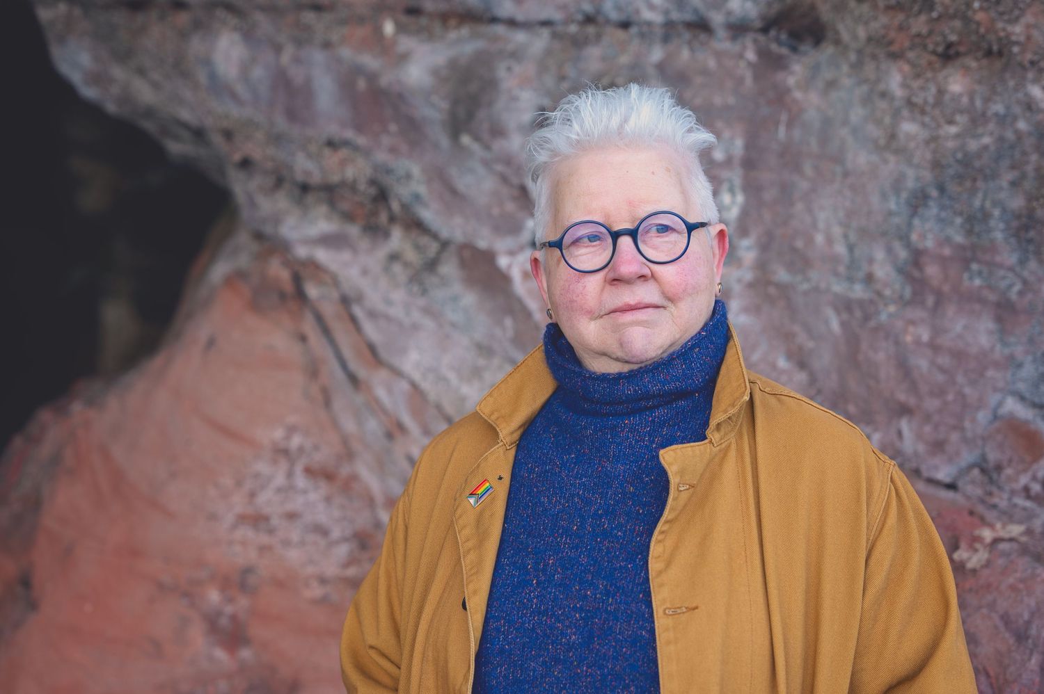 The Author Val Mcdermid with short white hair and round glasses wearing a mustard jacket and blue turtleneck sweater in the Weymss caves