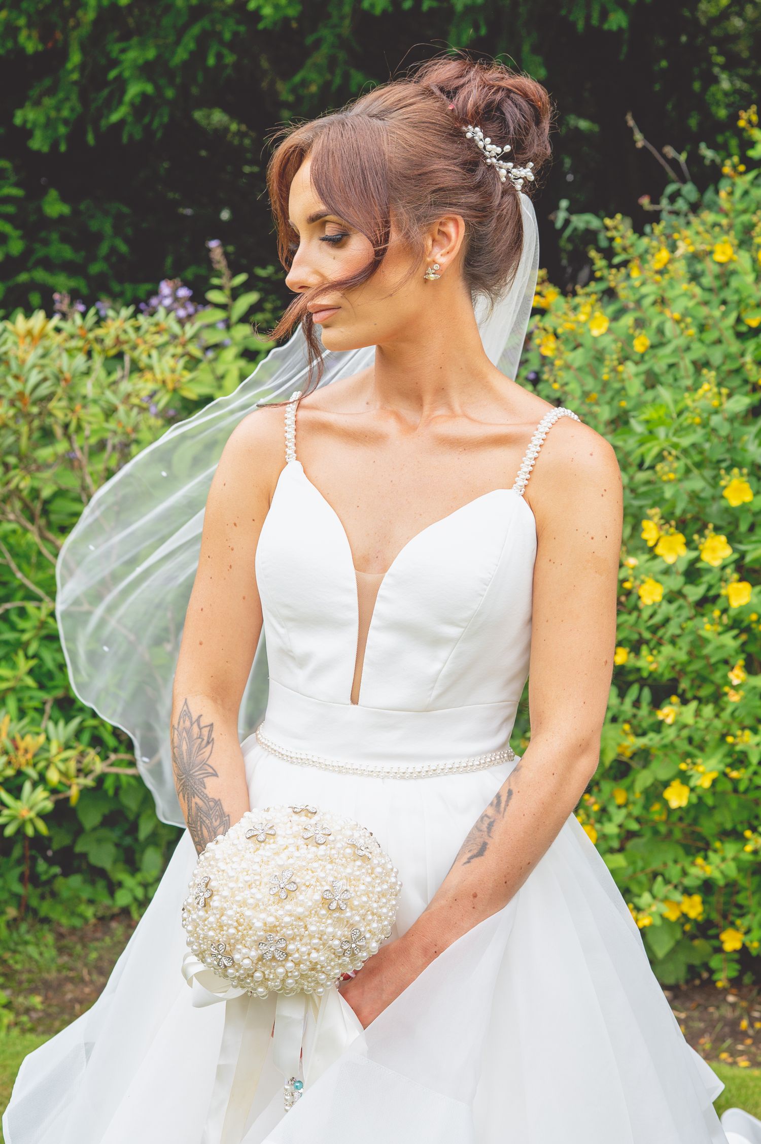Bride in white plunging neckline dress with veil and crystal hairpiece holds bouquet in garden setting.