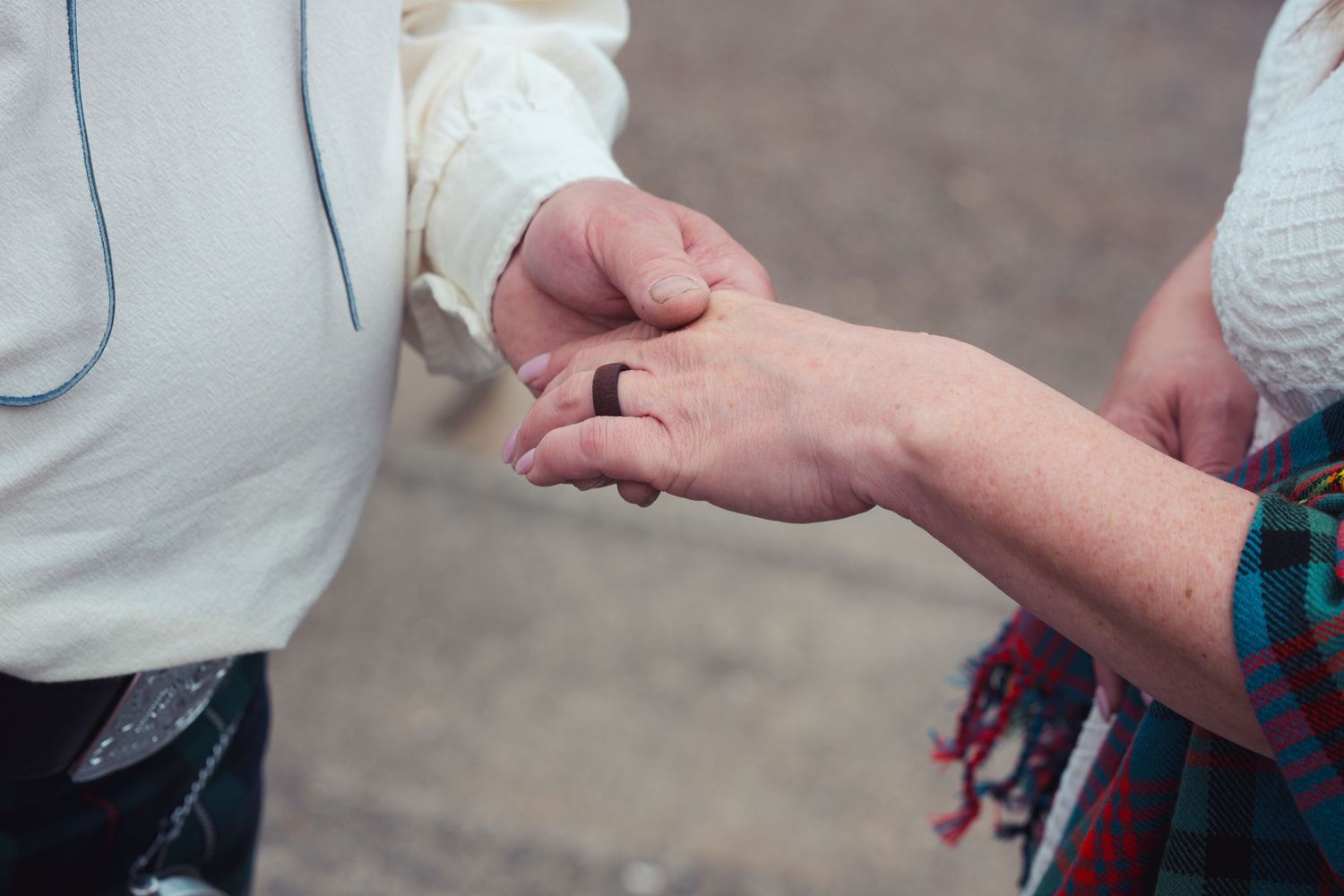 Close-up of two people holding hands, one wearing a dark wedding ring, dressed in traditional Scottish attire.
