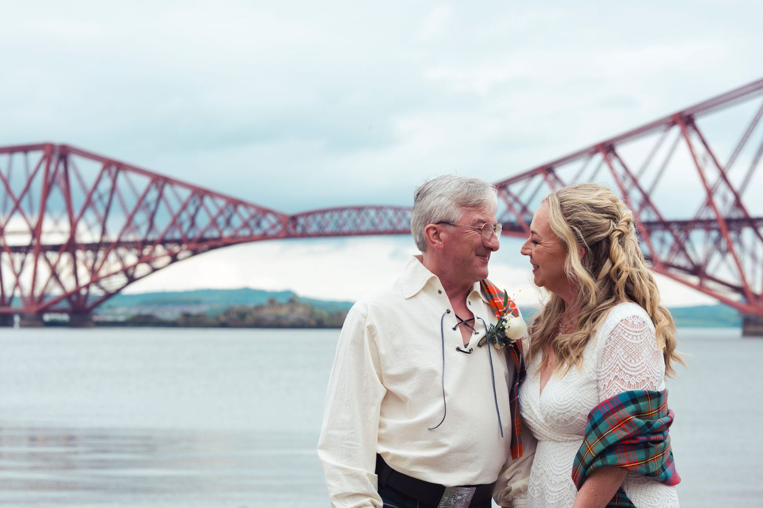 Couple smiling at each other near the Forth Bridge in Scotland, bride in lace dress with tartan shawl, groom in white shirt.