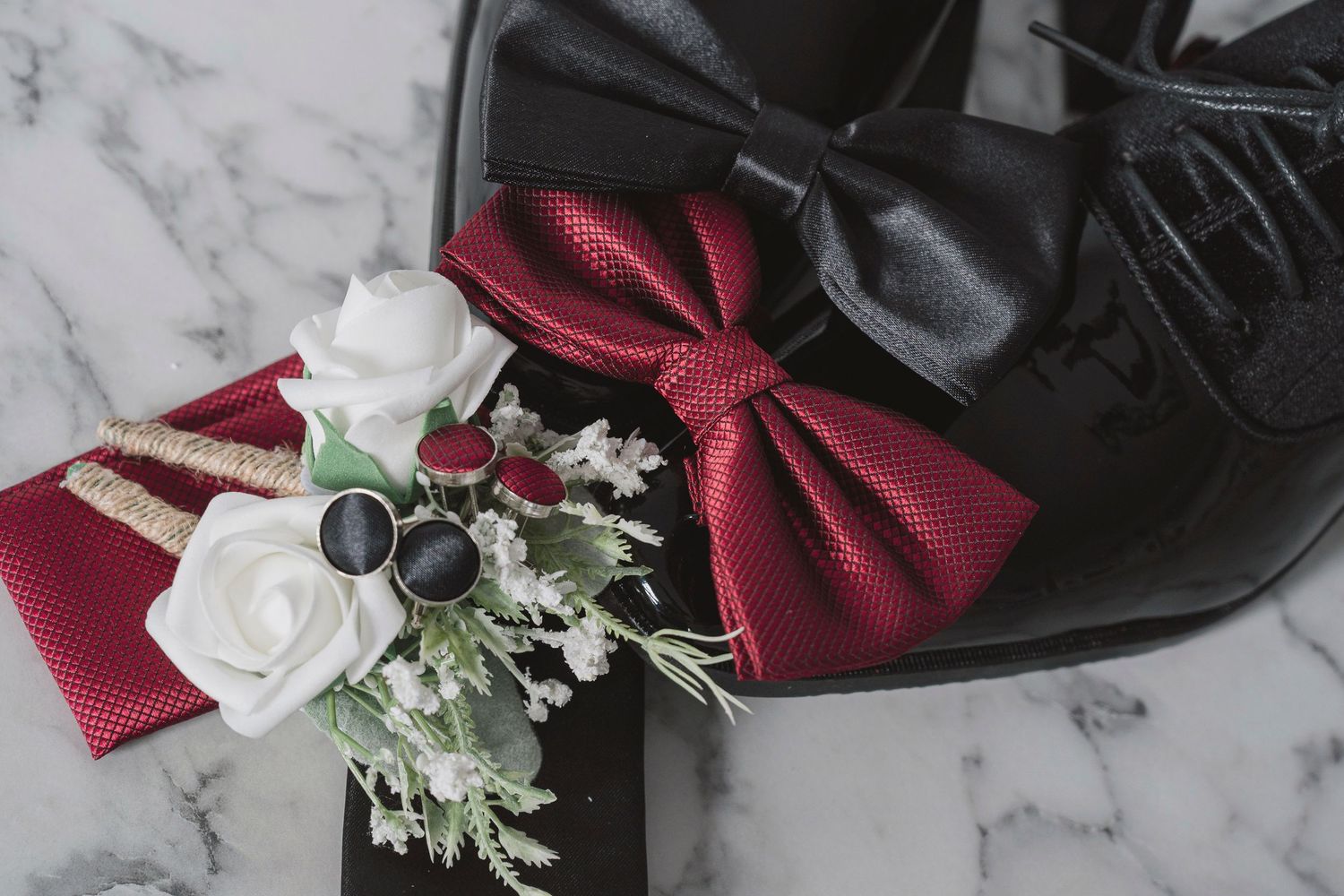 Elegant wedding accessories featuring a burgundy bow tie, white floral boutonniere, cufflinks, and black dress shoes on marble.
