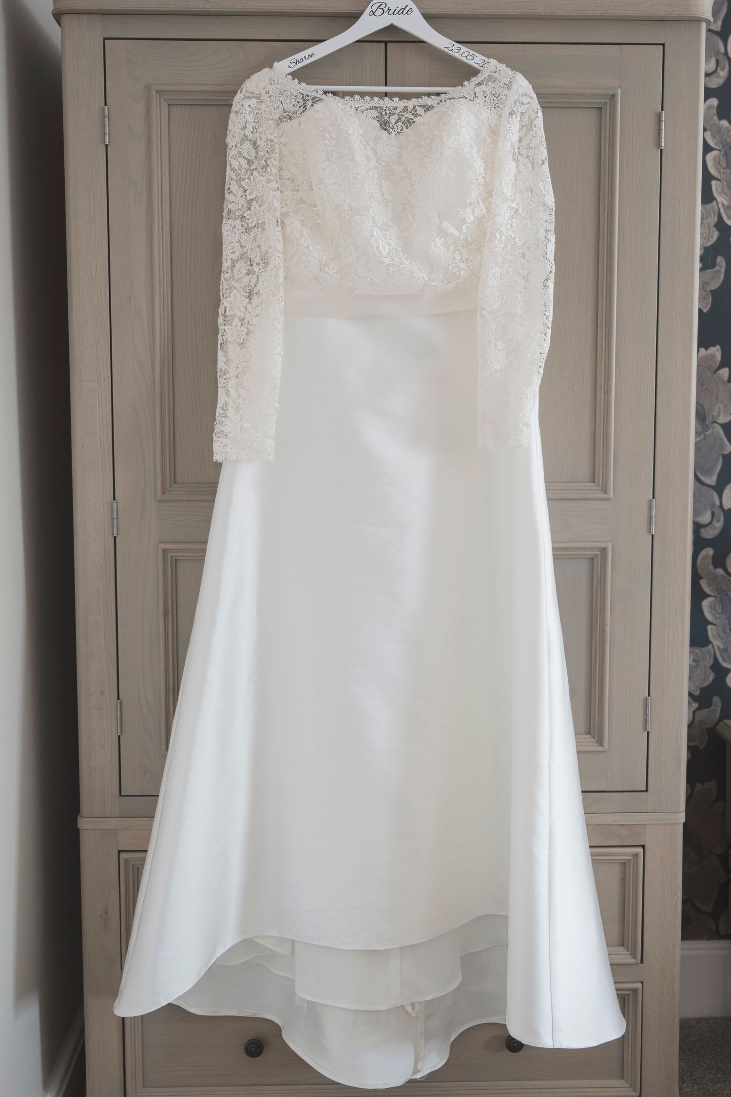 Elegant white wedding dress with lace long sleeves and satin skirt hanging on a wardrobe with a Bride hanger.