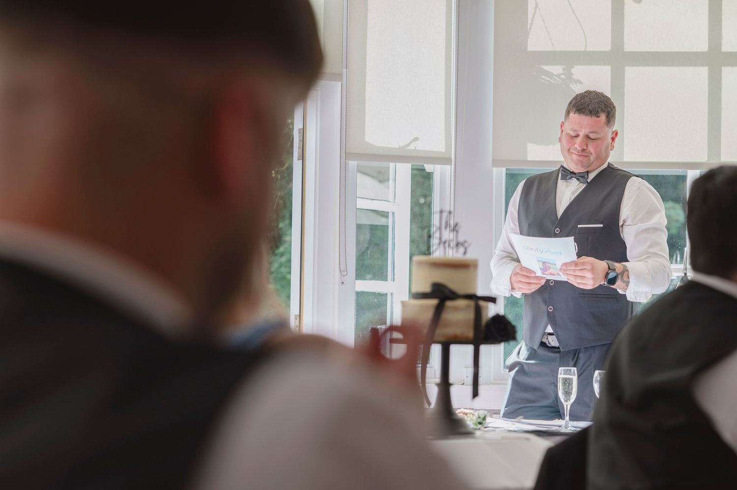 Man in vest and bow tie delivers a speech at an elegant wedding reception while guests listen attentively.