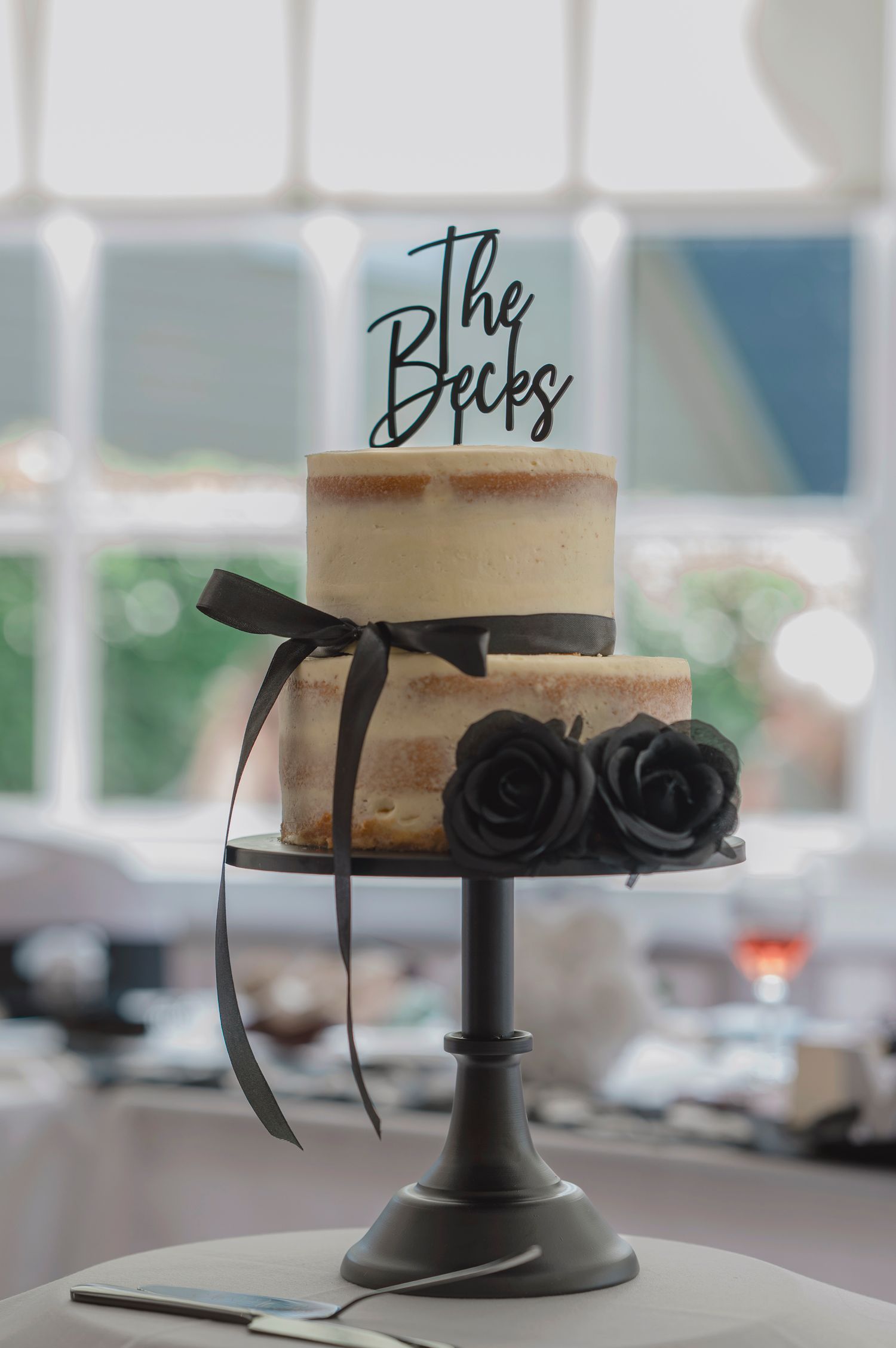 Two-tier semi-naked wedding cake with black roses, ribbon, and The Becks cake topper on a dark cake stand.