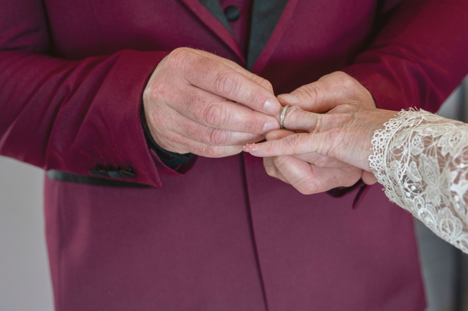 Person in burgundy suit placing a wedding ring on another persons hand during a marriage ceremony.