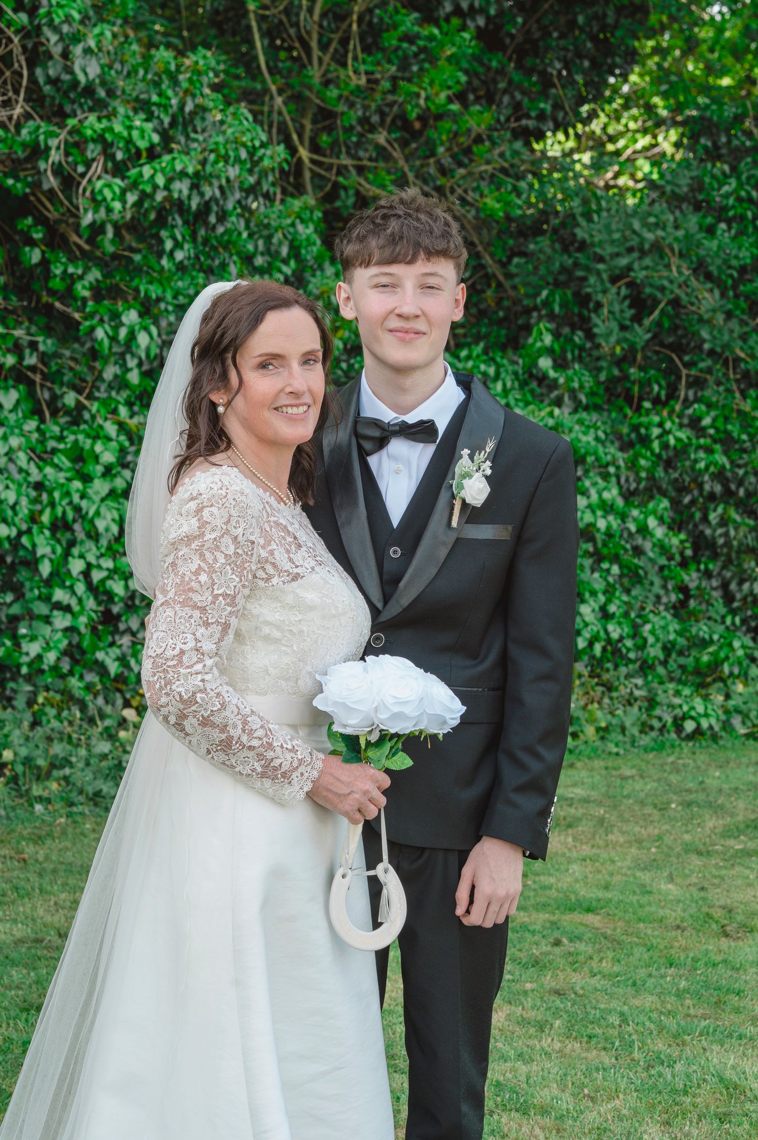 Bride in lace wedding gown holding white flowers poses with young man in black tuxedo outdoors.