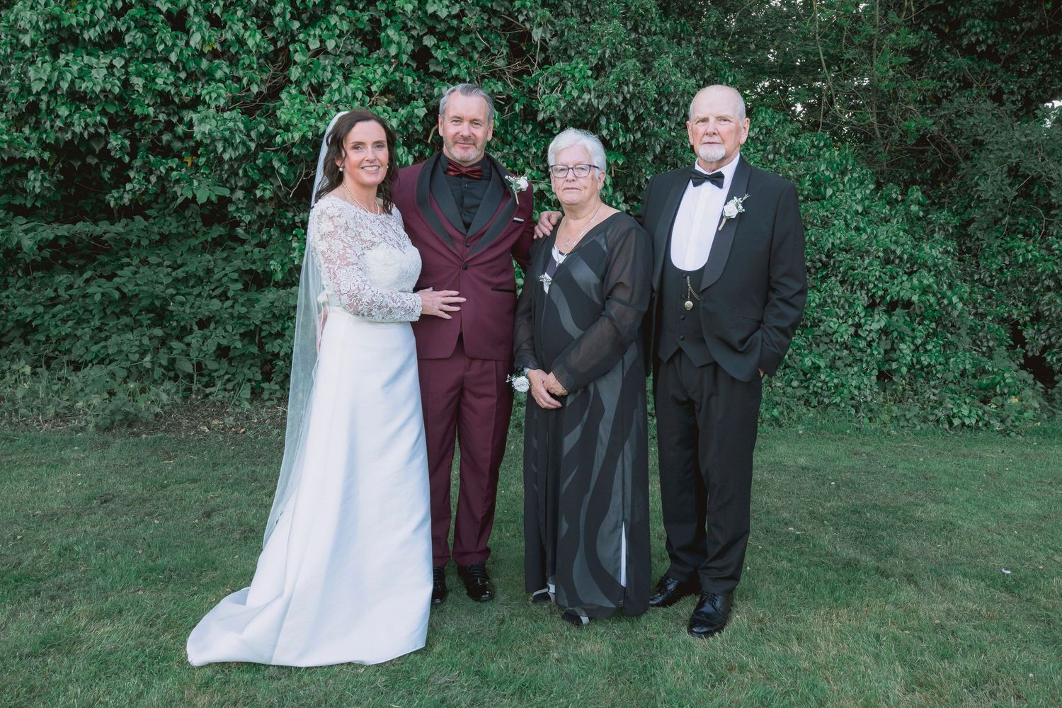 Wedding party of four posing outdoors, bride in white gown, groom in burgundy suit, couple in formal attire.