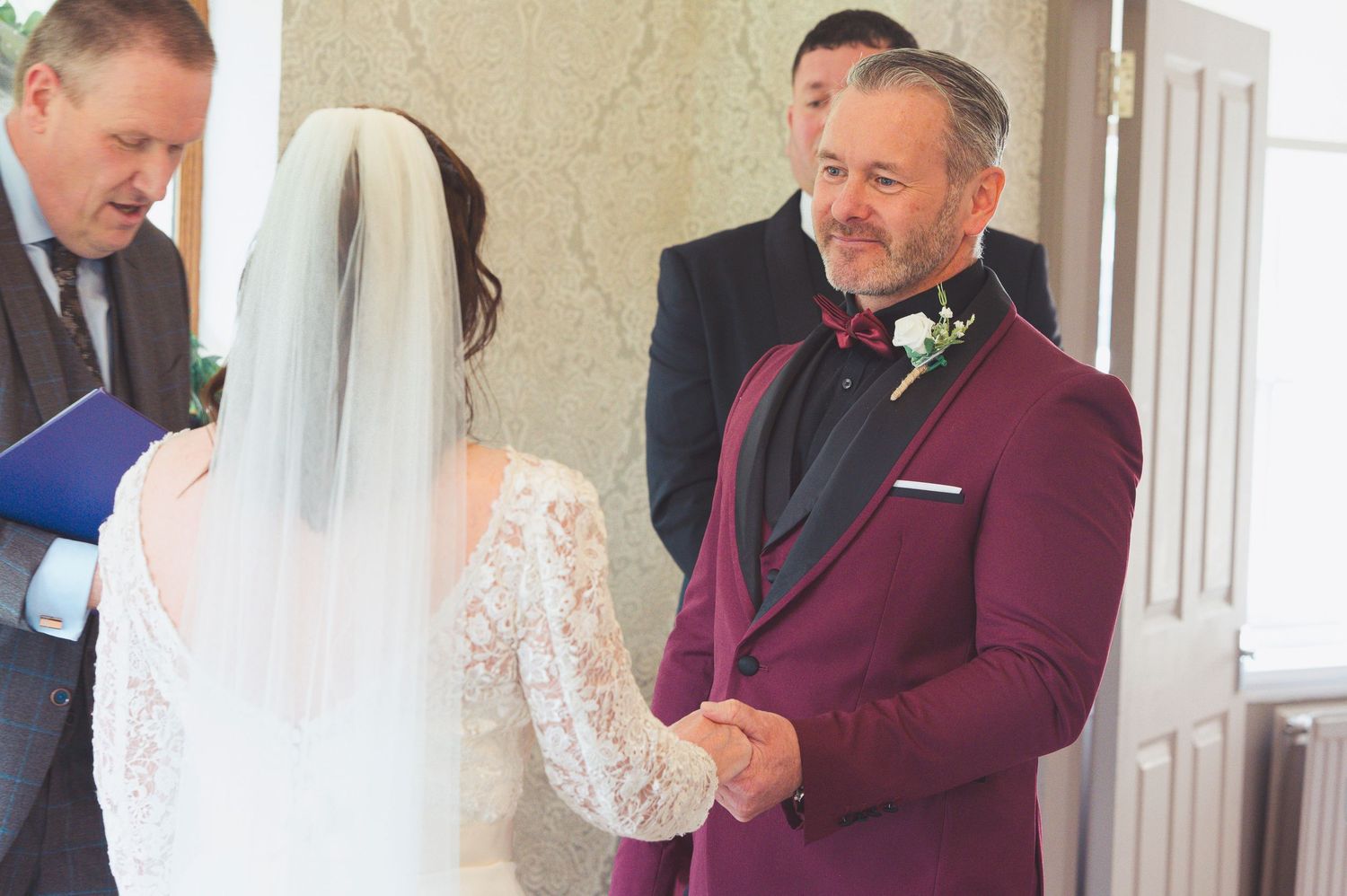 Groom in burgundy tuxedo holds bride's hands during outdoor wedding ceremony with officiant in background.