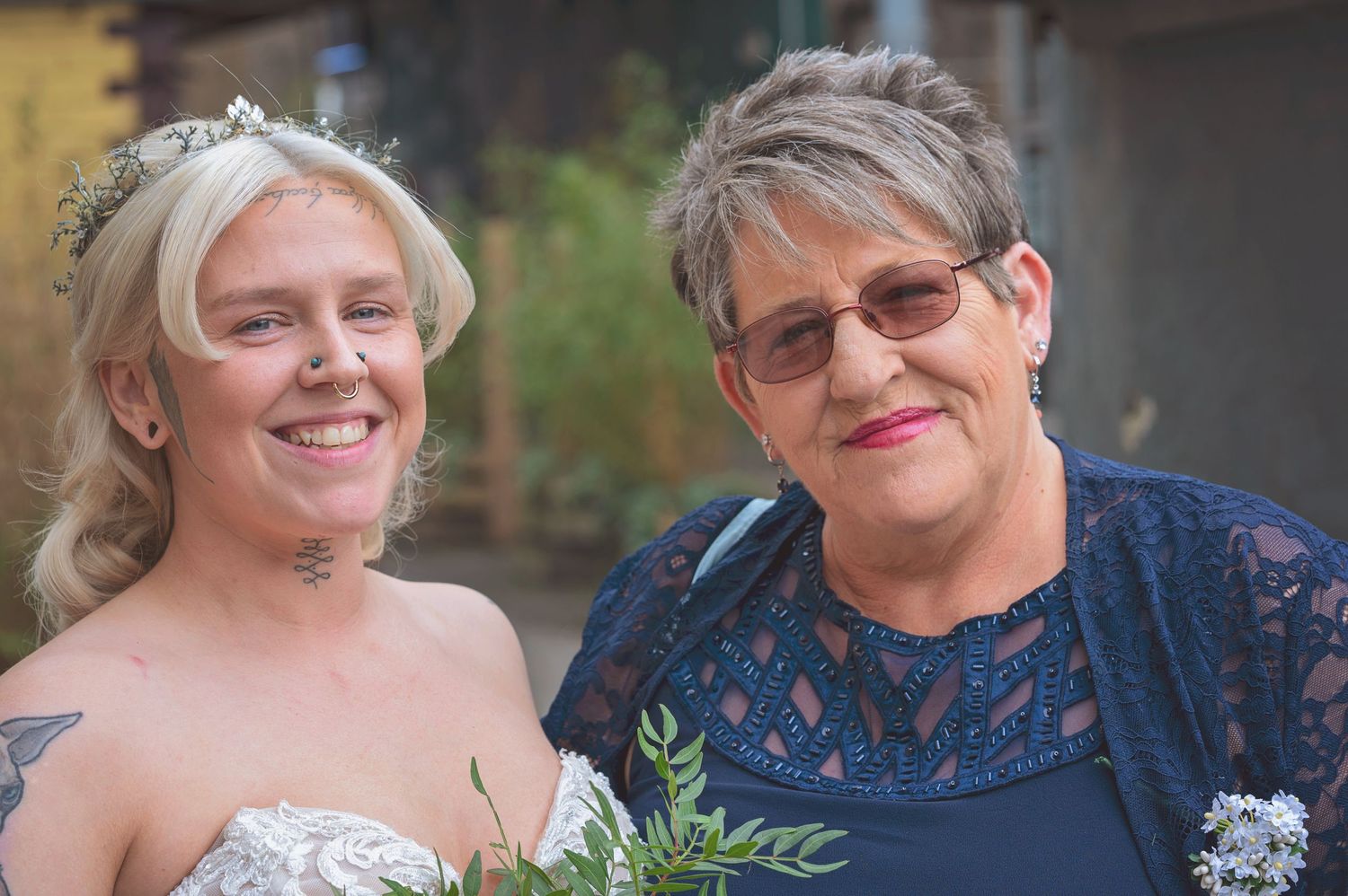 Smiling tattooed bride in white lace dress posing with older woman in blue at wedding.