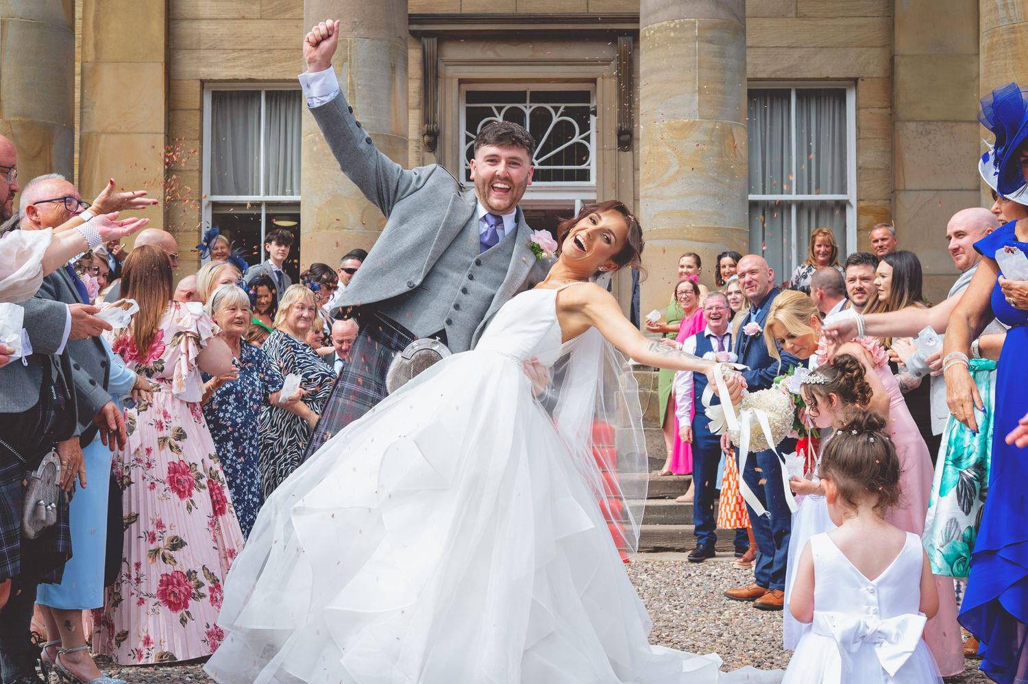 Wedding celebration outside a stone building with guests throwing confetti.