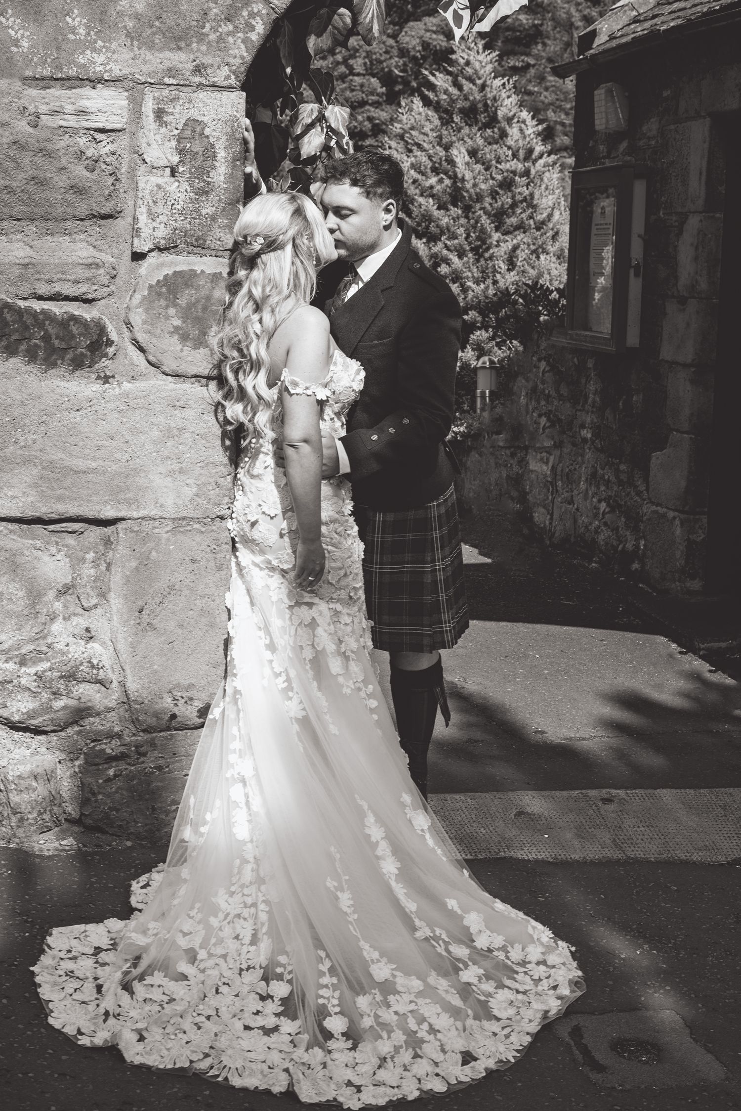 Black and white portrait of a couple in wedding attire with traditional Scottish kilt against stone wall.