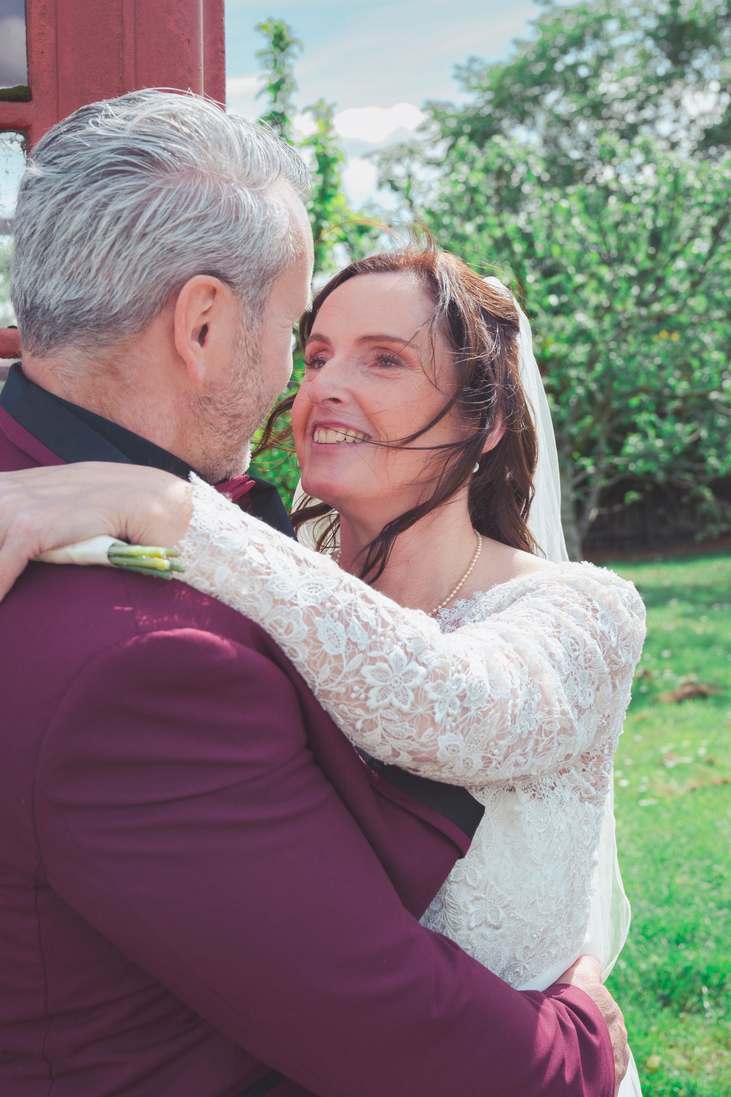Bride in lace dress embraces groom in burgundy suit, smiling joyfully outdoors on their wedding day.