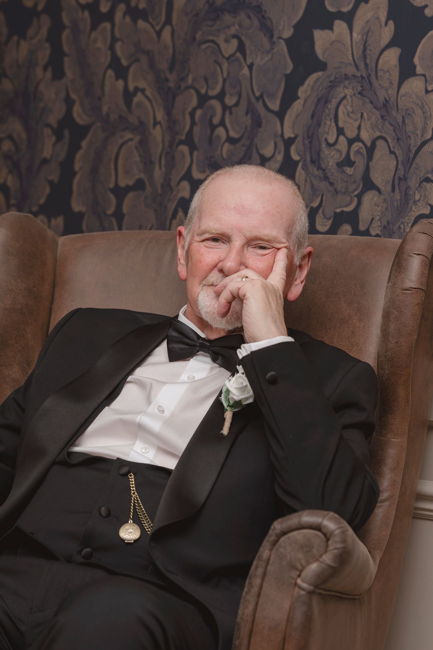 Elderly man in black tuxedo with bow tie and pocket watch sits relaxed in leather chair against ornate wallpaper.