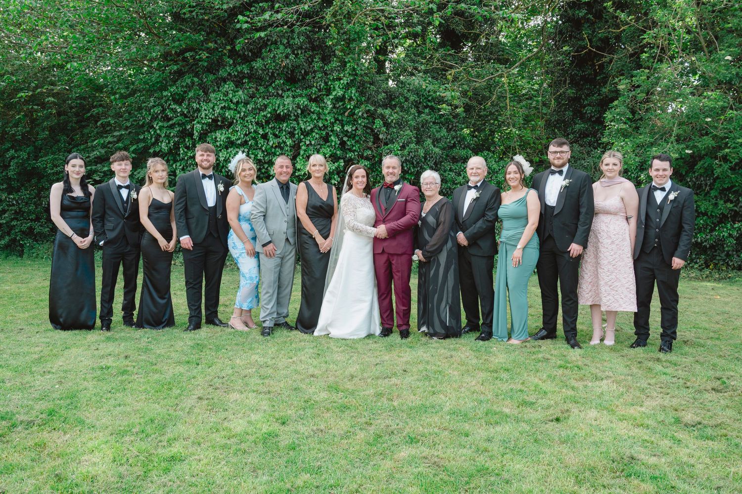 Wedding party poses outdoors on green lawn with trees, bride in white gown and groom in burgundy suit surrounded by guests.