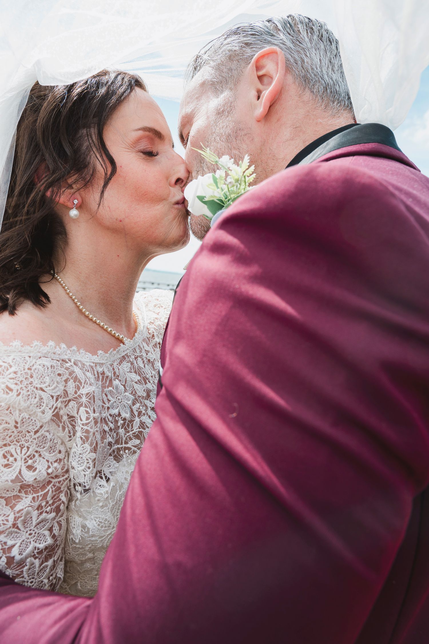 Bride and groom sharing a romantic kiss under a veil, groom in burgundy suit with boutonniere.