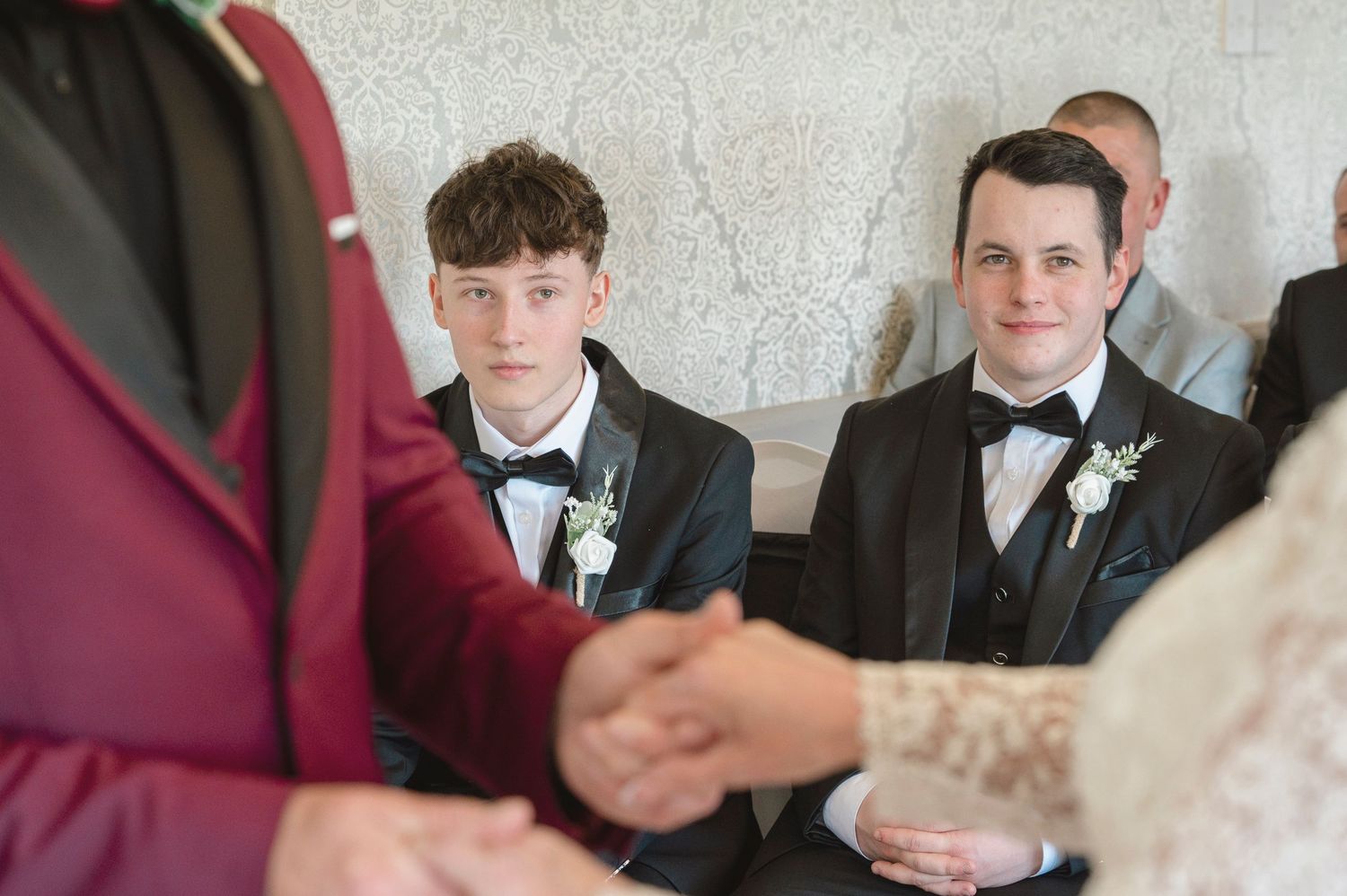 Two young groomsmen in tuxedos with boutonnieres watch a wedding ceremony, smiling warmly at the couple.
