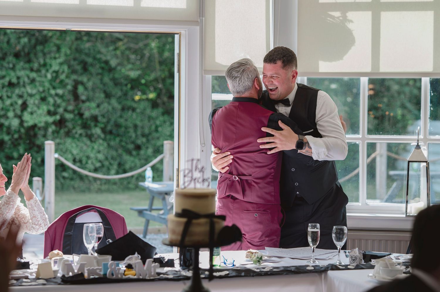Two men share a warm hug at a wedding reception, dressed in formal attire near an elegantly set dining table.