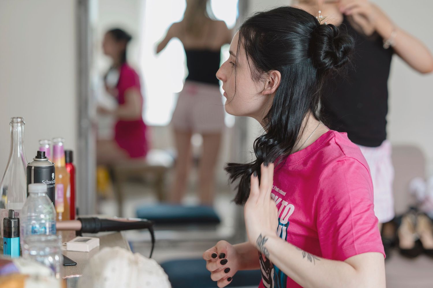 Woman in pink shirt braiding hair at vanity mirror with beauty products in dance studio.