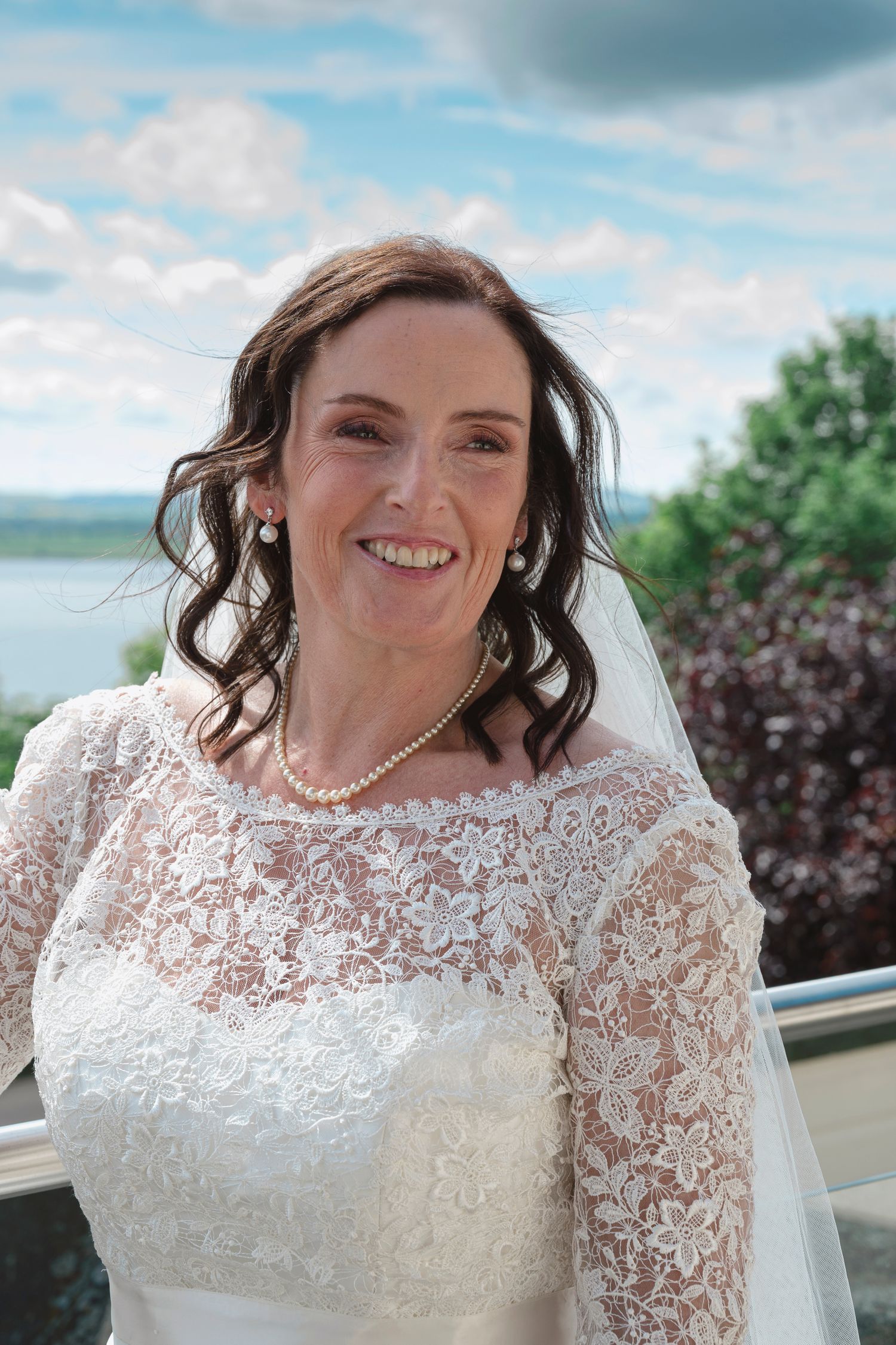 Smiling bride in lace wedding dress with pearl jewelry and veil, posing outdoors with blue sky backdrop.