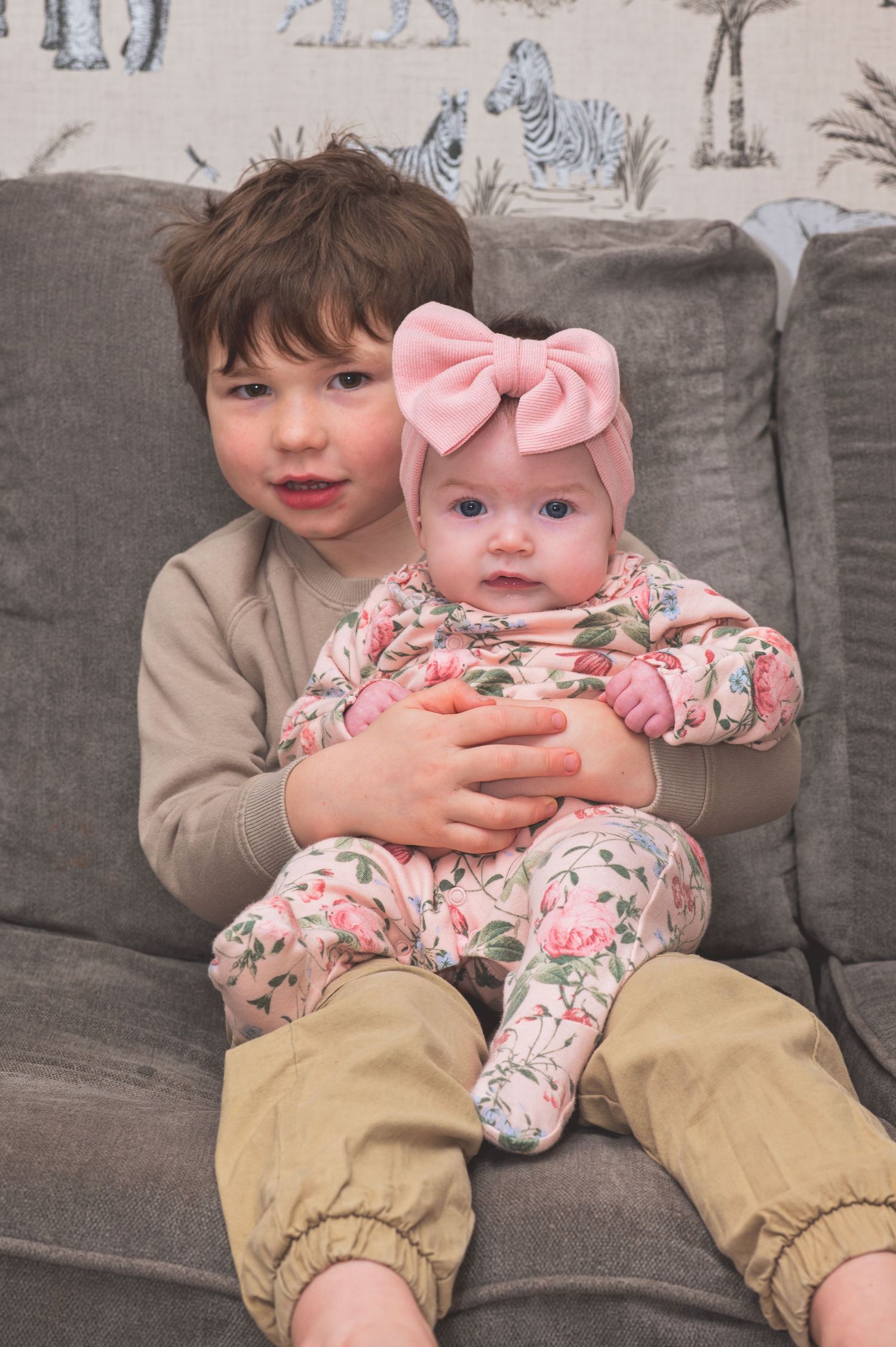 Two children sit together on a gray couch, one wearing khaki pants and the other in a floral pink outfit with bow.