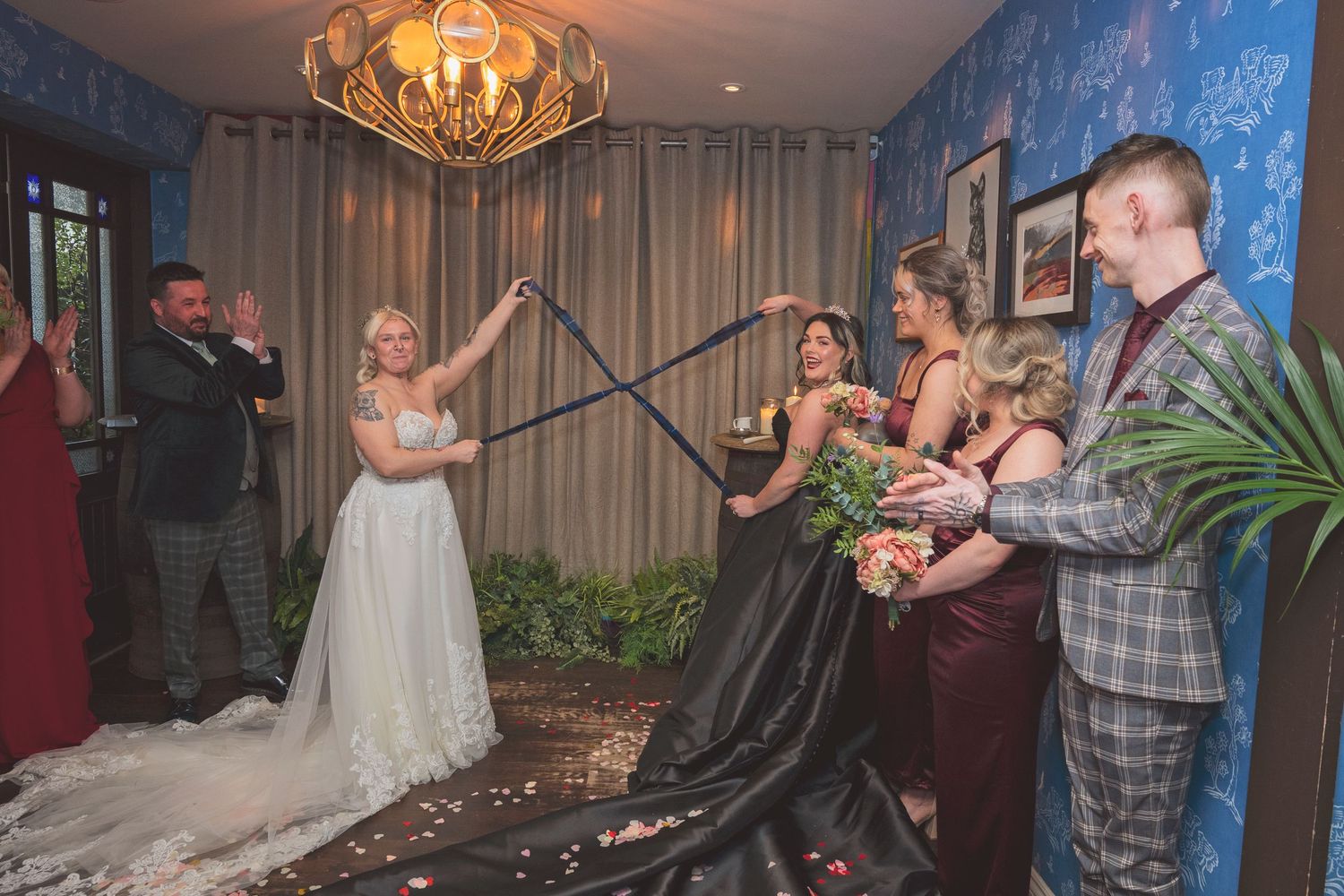 A bride in a white gown participates in a handfasting ceremony with wedding party in a decorated venue.
