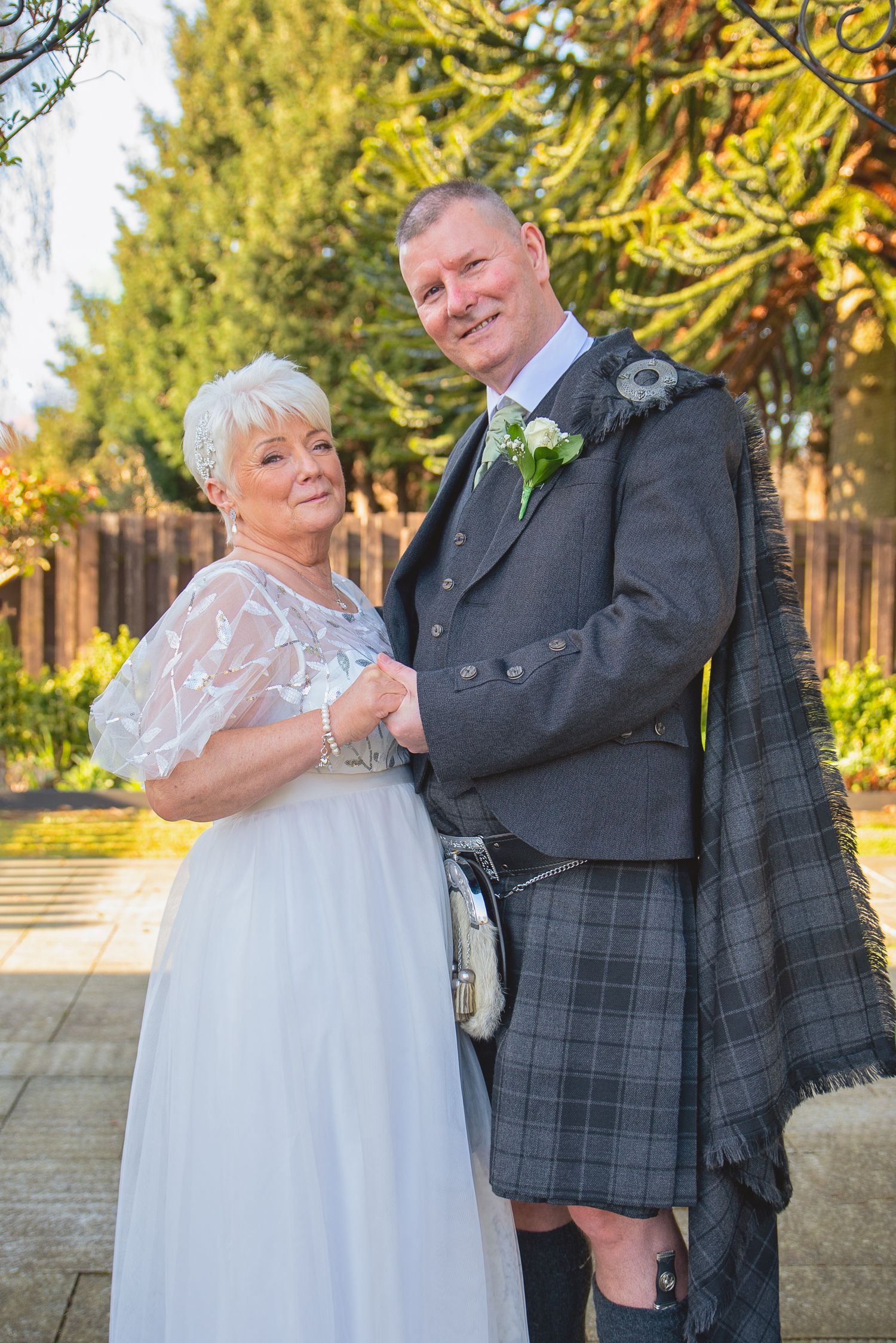 Couple in traditional Scottish wedding attire pose together in a garden setting.