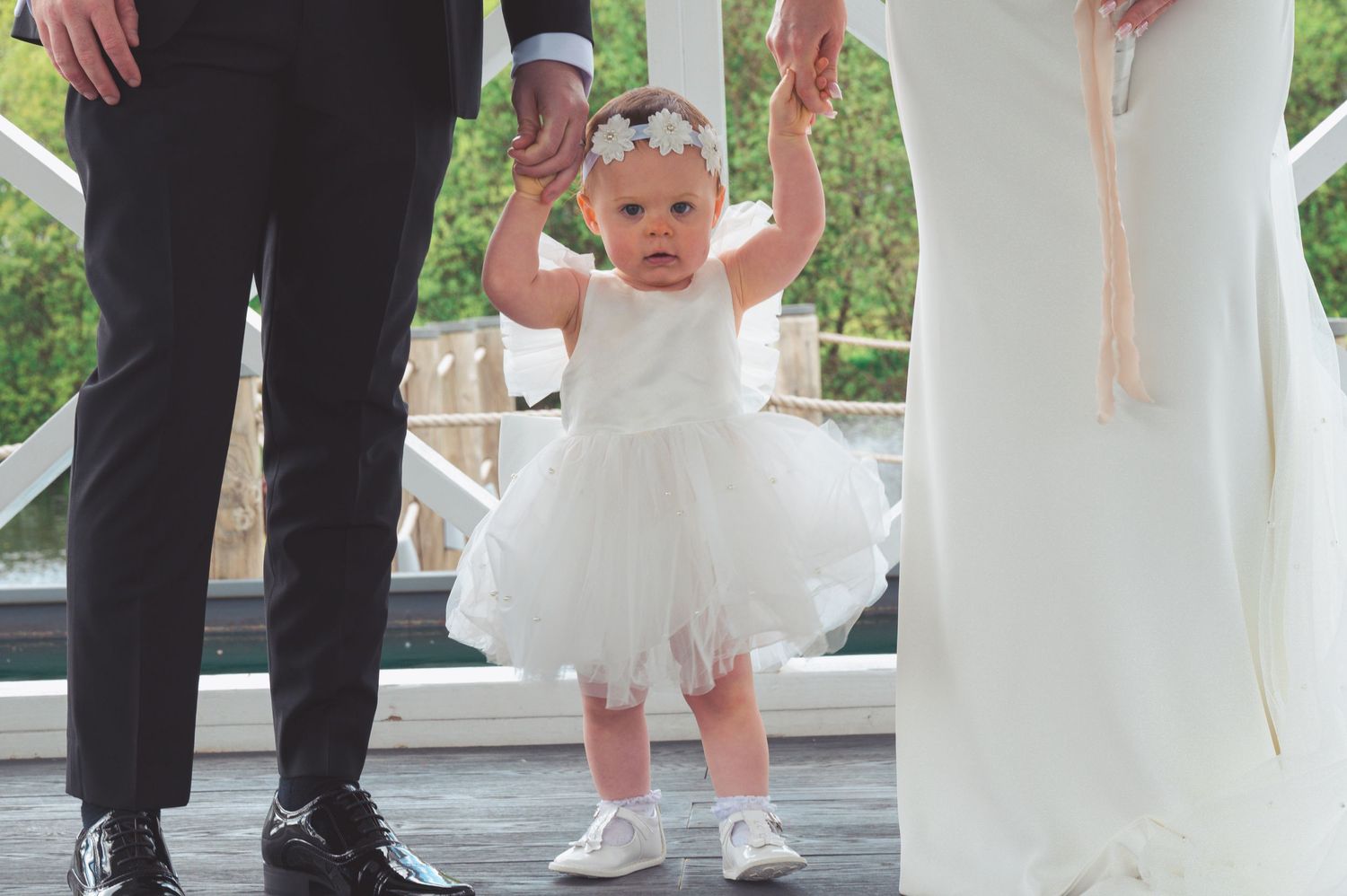Adorable baby girl in white tutu dress held by bride and groom at wedding ceremony.